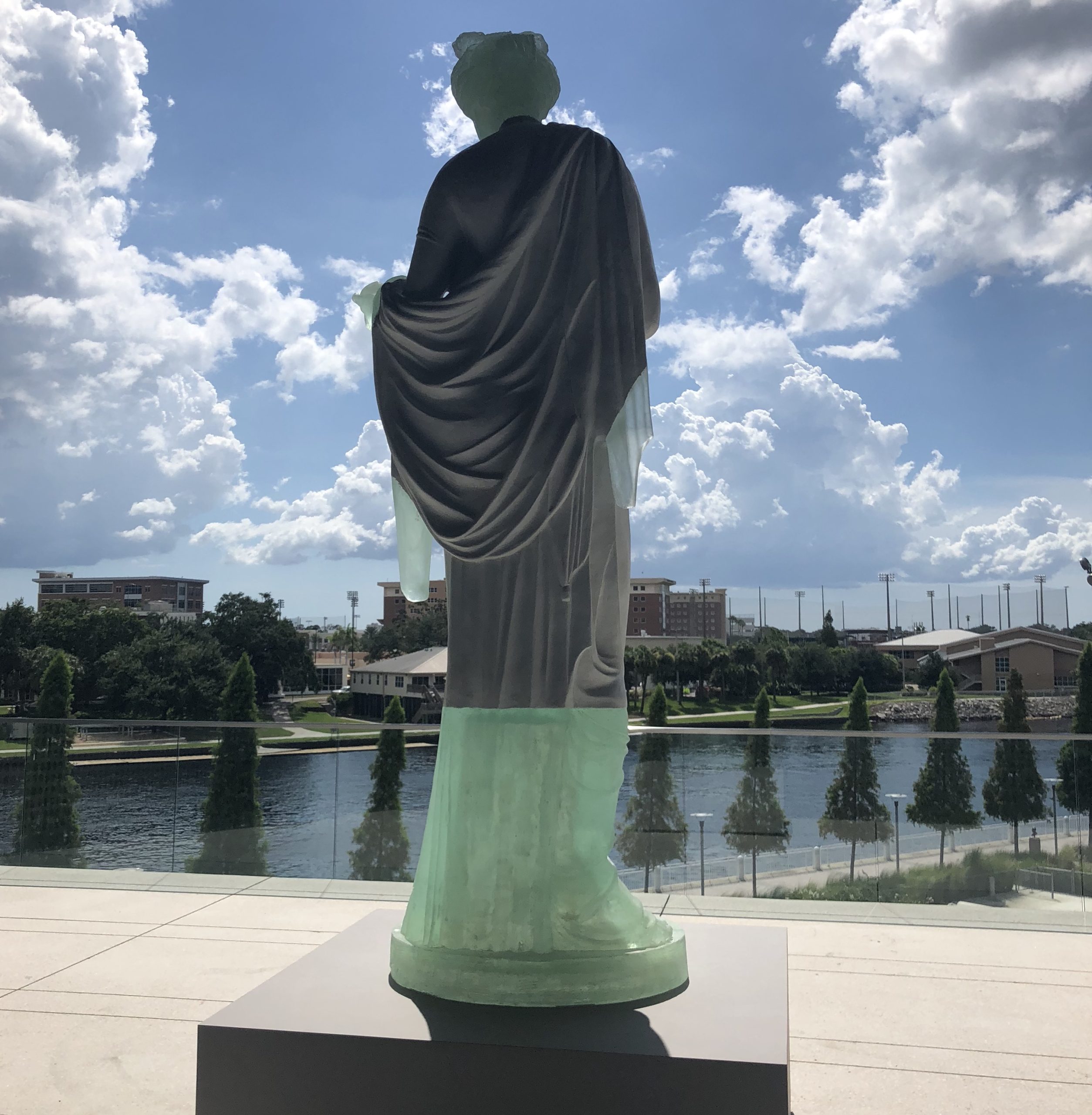 Freestanding sculpture of a woman in a toga, seen from behind, in front of a reflecting pool. Parts of the sculpture are dark and others a translucent green