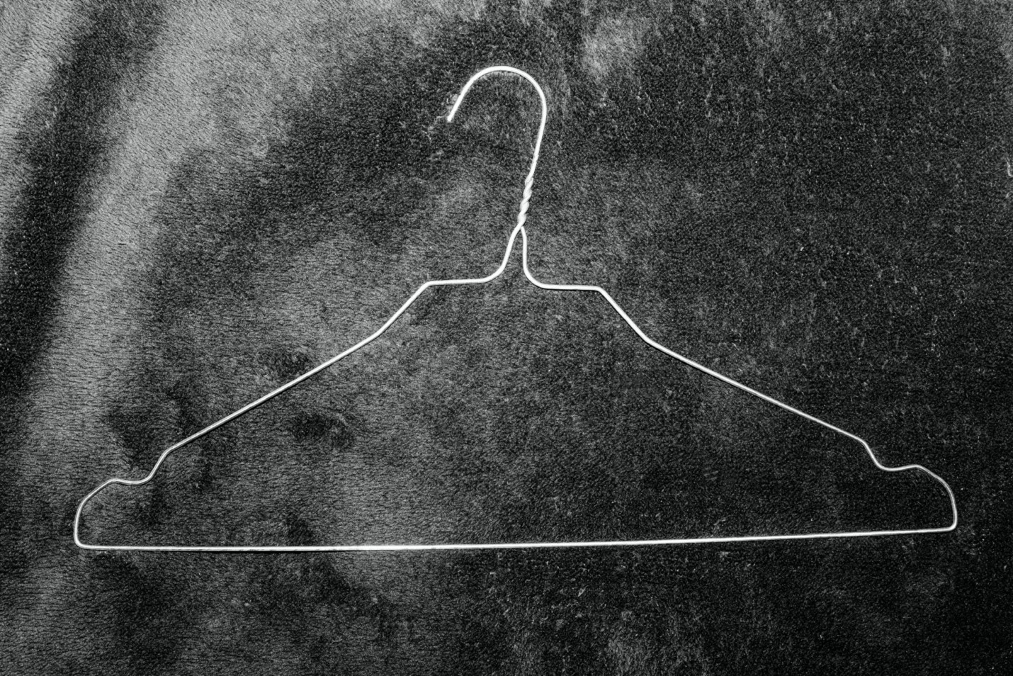 Black-and-white photographic image of a white wire coat hanger against a mottled dark background.