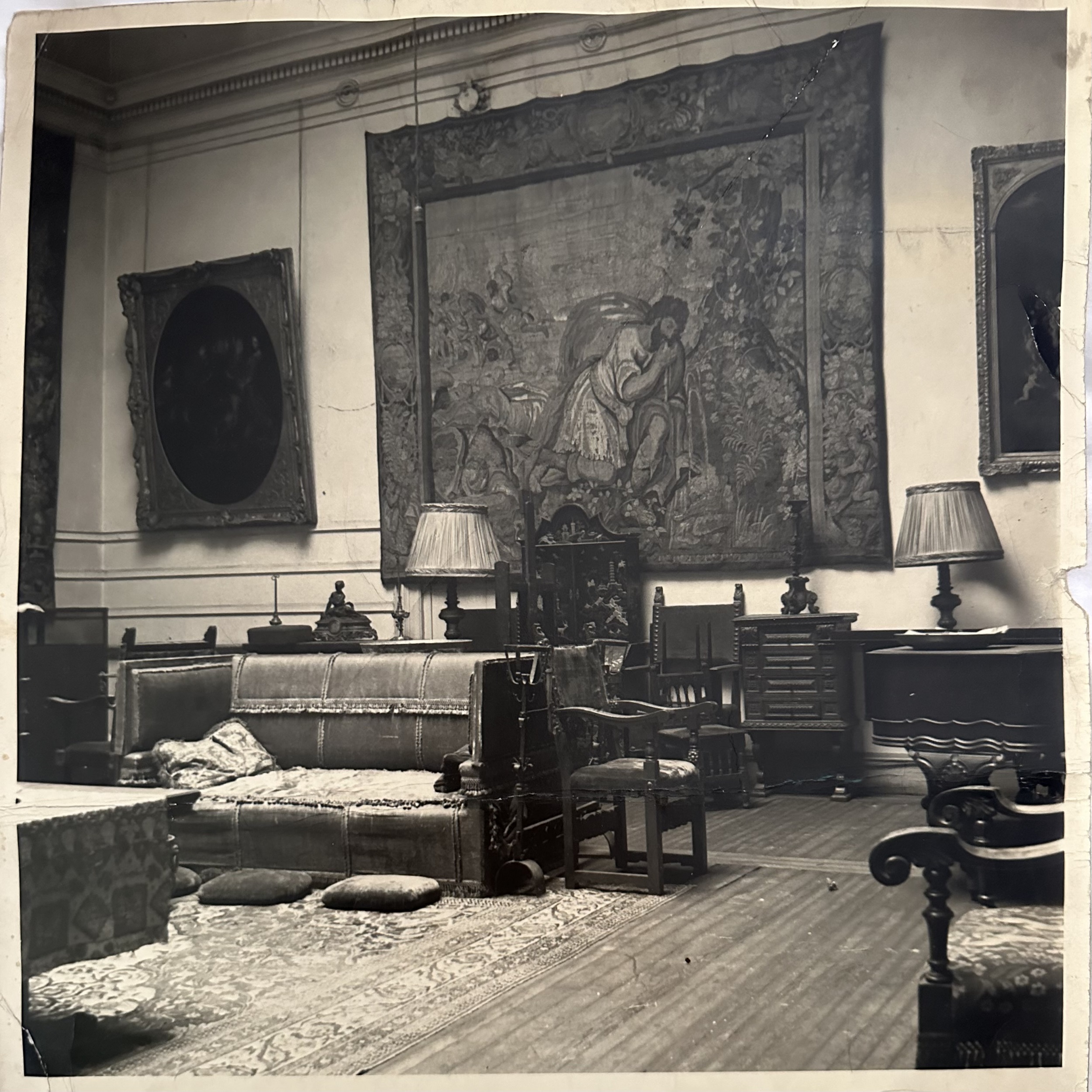 Black-and-white historic photograph of the interior of a high-ceilinged domestic space hung with paintings and a tapestry.