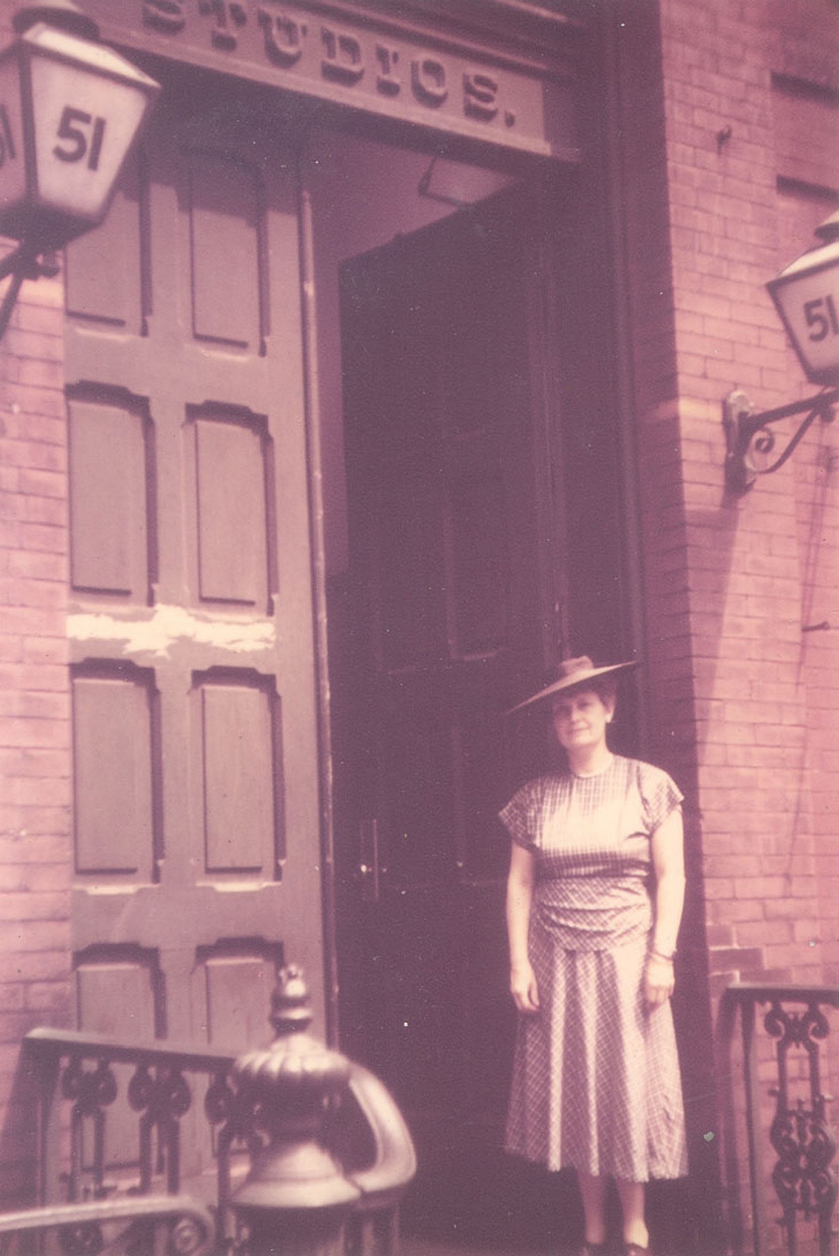 Purple-tinged photograph of a woman in a dress and hat standing in front of a large door, above which is the word "STUDIOS." A lantern in the upper left has the number "51."