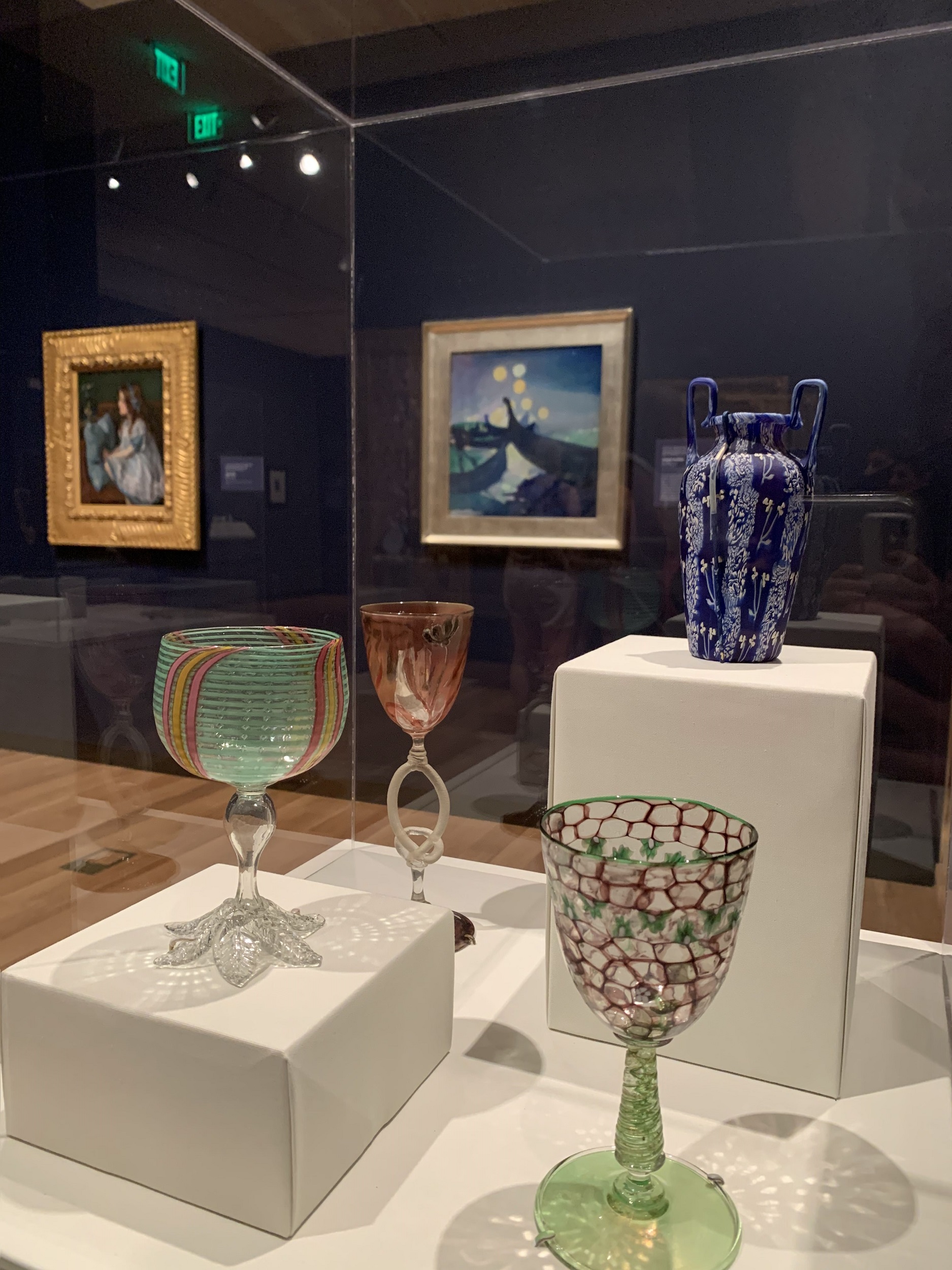 Interior view of a museum gallery, with a case containing several pieces of art glass in the foreground and paintings hung on a dark blue wall in the background.