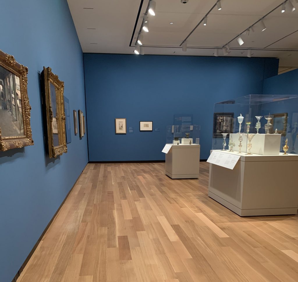 Interior view of a gallery with paintings hung on mid-blue walls. In the center of the room are a pair of large glass-topped cases containing several pieces of art glass.