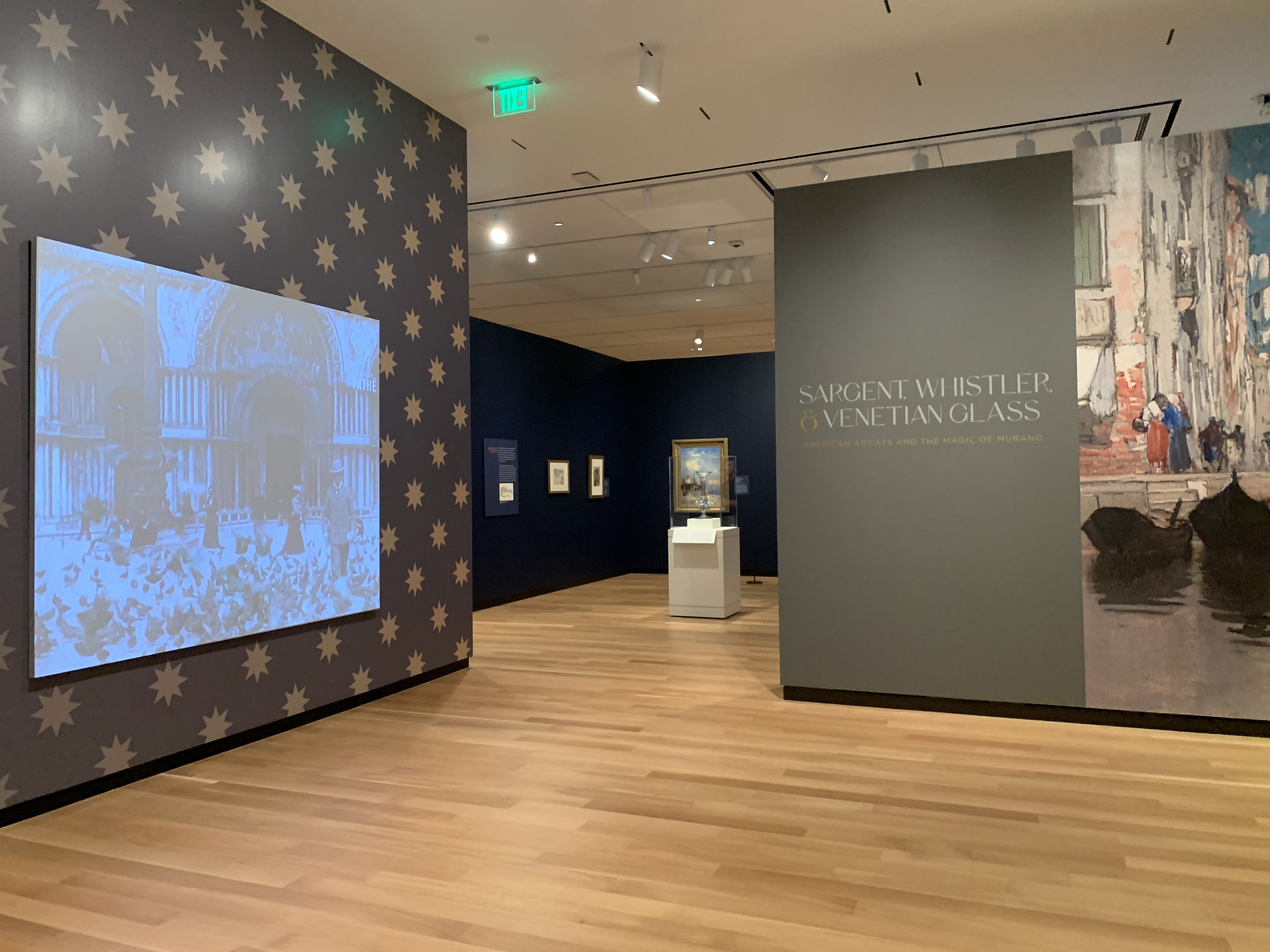 Entrance to a museum exhibition; the wall on the left has a digital projection of St. Mark's square, Venice, on a wall covered with stars on a blue ground. The wall on the right reads "SARGENT, WHISTLER & VENETIAN GLASS" and has a reproduction of a painting. 