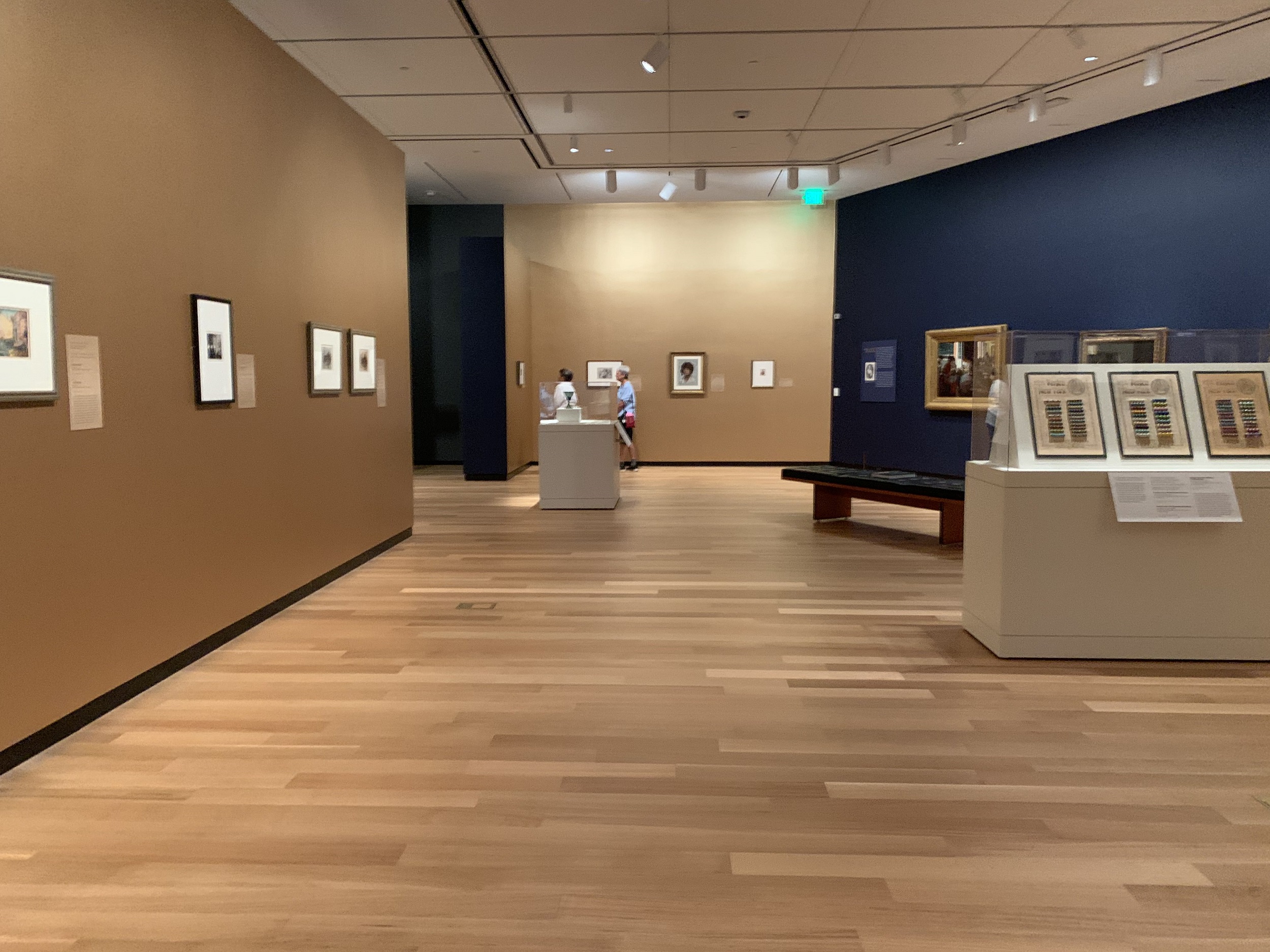 Interior of a museum gallery with works on paper hung on mustard-colored walls. There is a glass-topped case to the right.