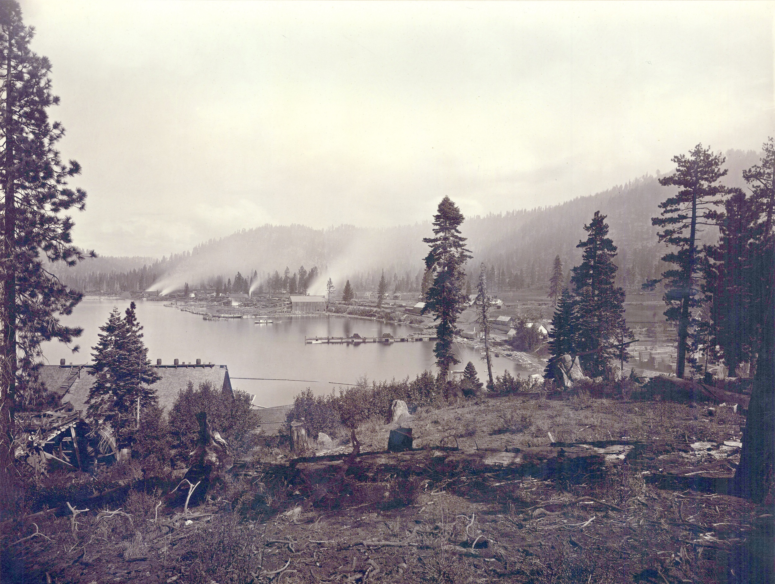 Black-and-white photograph of a lake surrounded by pine trees and rustic encampments.