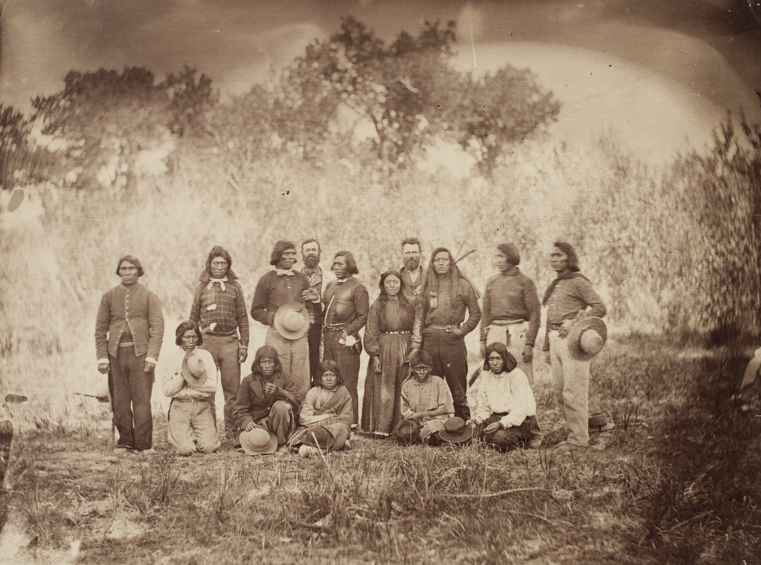 Sepia-toned group photograph of thirteen Native Americans and two Euro-American men in a grassy field, with trees behind them