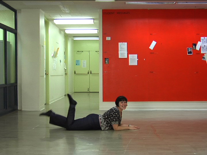 Photograph of a woman in black pants lying on her stomach on a terrazzo floor in front of a red wall, kicking her heels up and smiling.