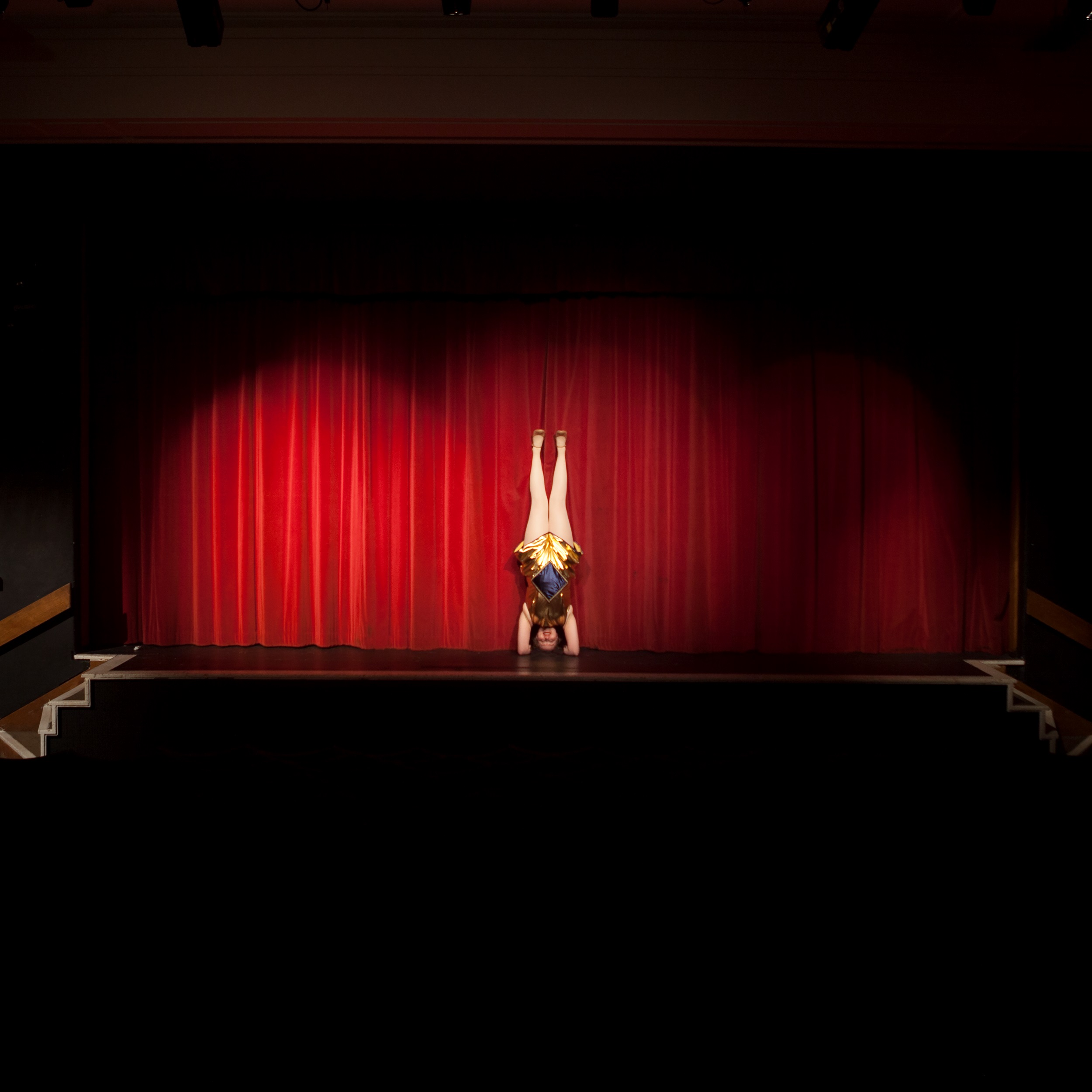 Photograph of a woman in a short blue-and-gold costume doing a headstand on an empty stage, in front of a spotlight red curtain.