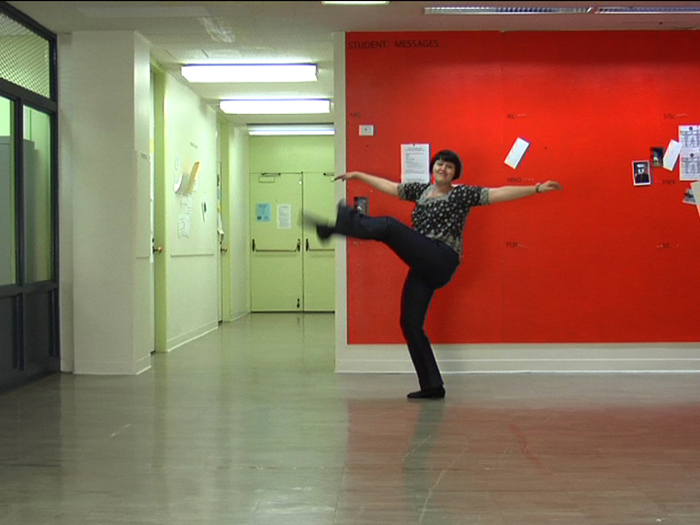 Photograph of a woman in black pants doing a high kick on a terrazzo floor in front of a red wall