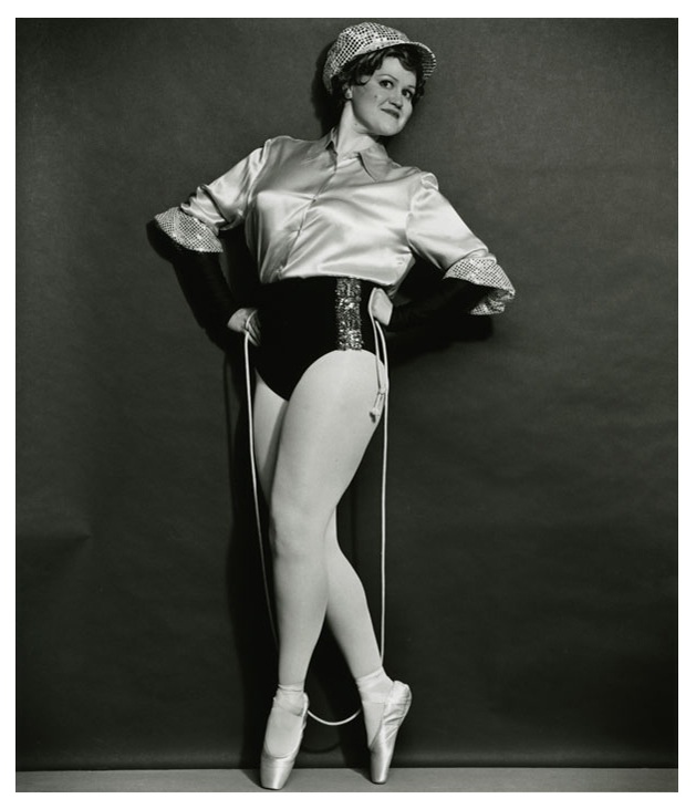 Black-and-white photograph of a showgirl wearing black briefs, a long-sleeved satin shirt, and toe shoes