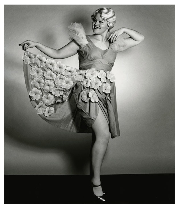 Black-and-white photograph of a costumed showgirl wearing a flower-adorned skirt that she holds out with one arm