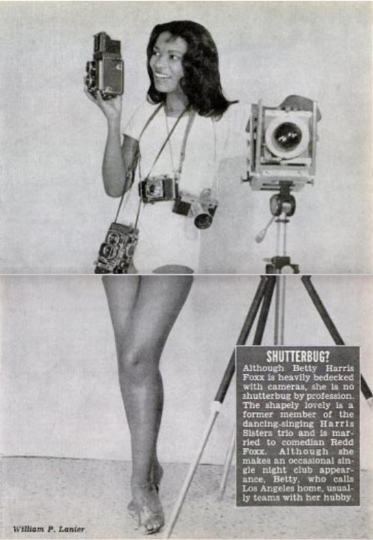 Black-and-white halftone image of a Black woman wearing a short white romper, standing next to a camera on a tripod, holding another camera, and wearing several others around her neck. A text panel at the lower left reads "Shutterbug: Although Betty Harris Foxx is heavily bedecked with cameras, she is no shutterbug by profession. The shapely lovely is a former member of the dancing-singing Harris Sisters trio and is married to comedian Redd Foxx. Although she makes an occasional single night club appearance, Betty, who calls Los Angeles home, usually teams with her hubby.