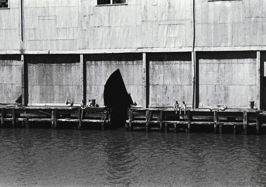 Black and white photograph of a corrugated metal building on an industrial pier with a triangular-shaped cutout at its base. Several nude men sit and recline on the dock outside.
