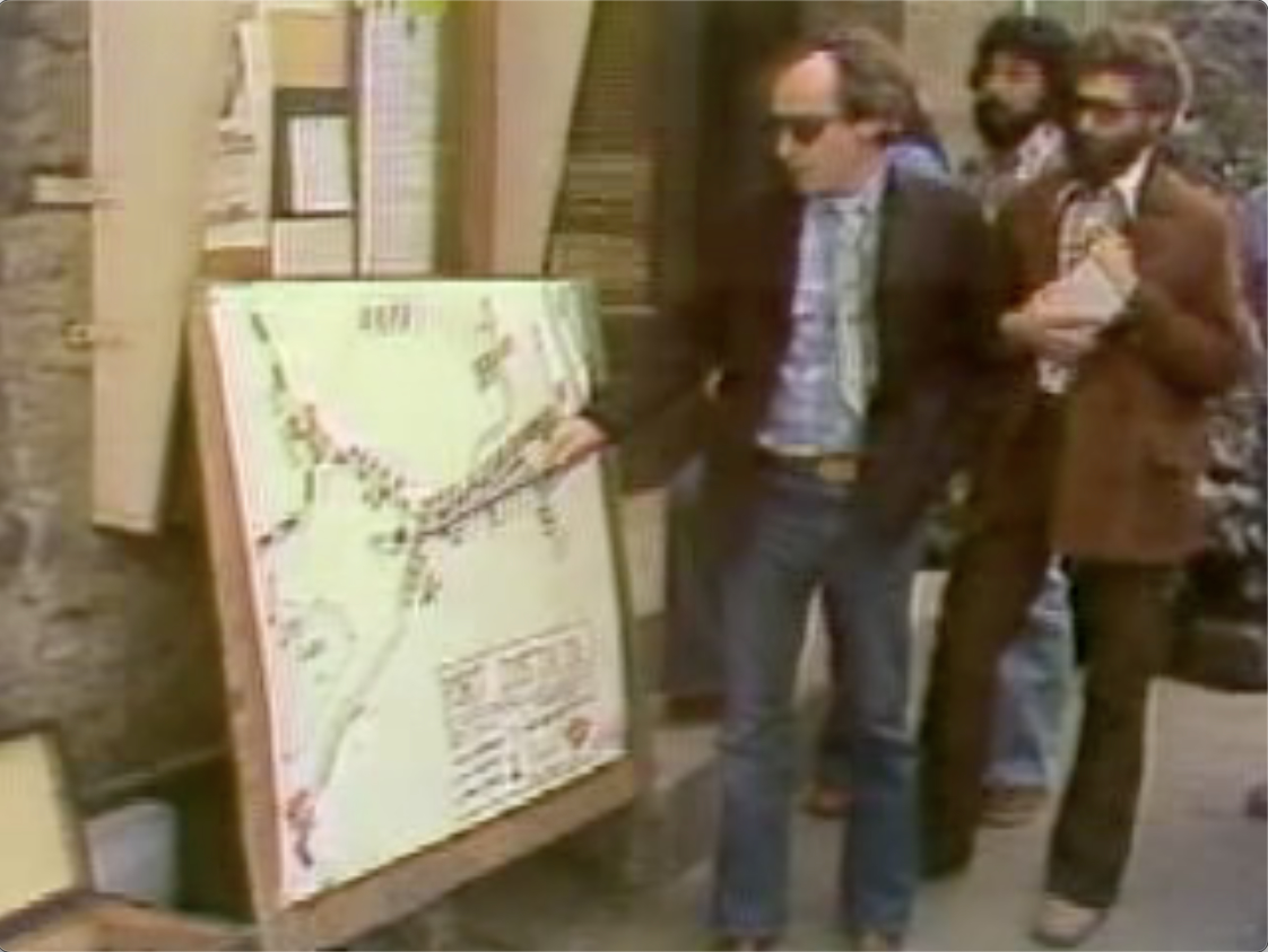 Color video still of three men in standing to the right of a map placed on a sandwich board. One man, wearing a suitcoat and sunglasses, is using a pointer to indicate something on the map.