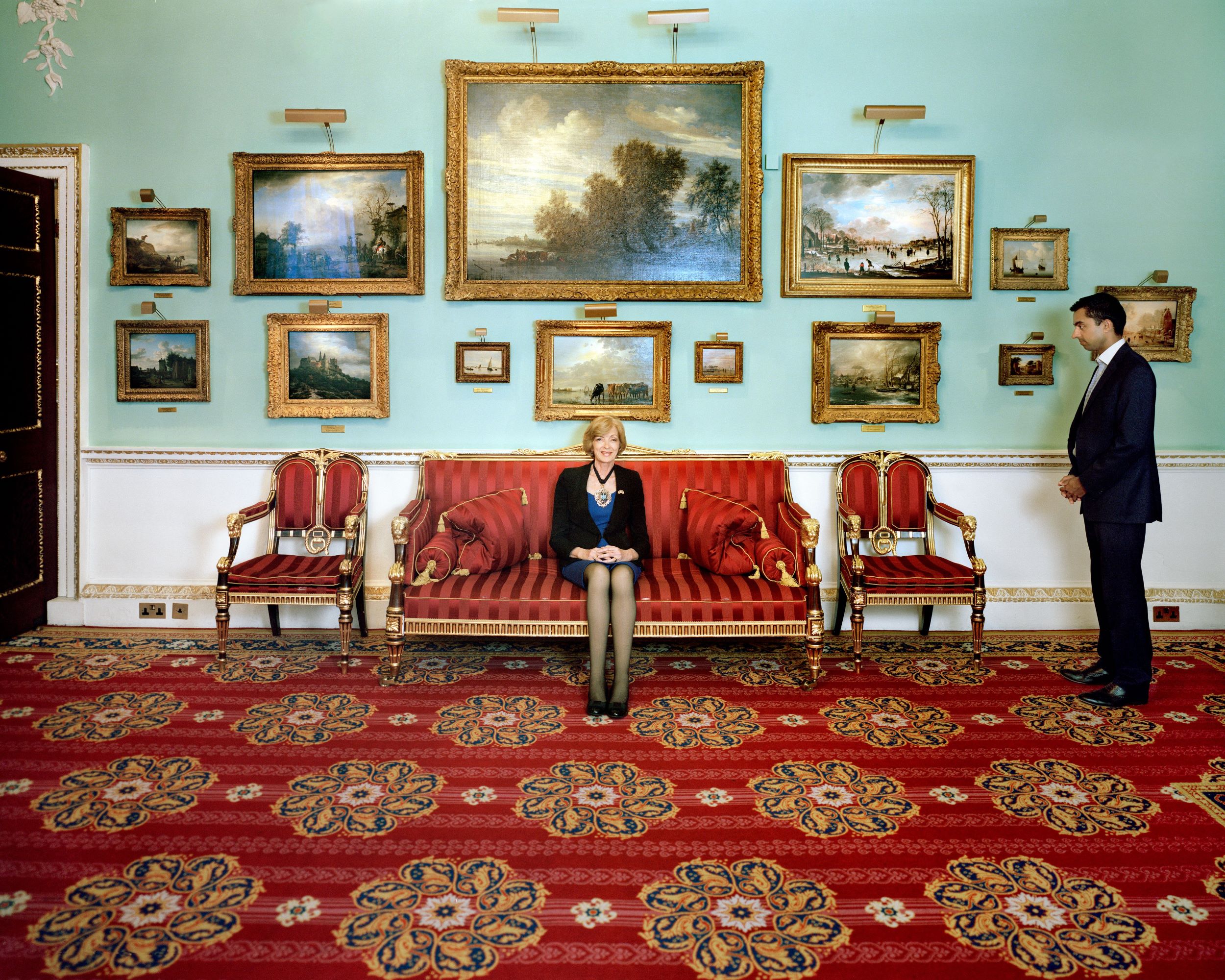 Color photograph of a blond woman in a black suit sitting on a red Empire-style couch in room with an ornate red carpet and multiple landscape paintings on the walls. To the right is a man in a dark suit turned toward the woman.