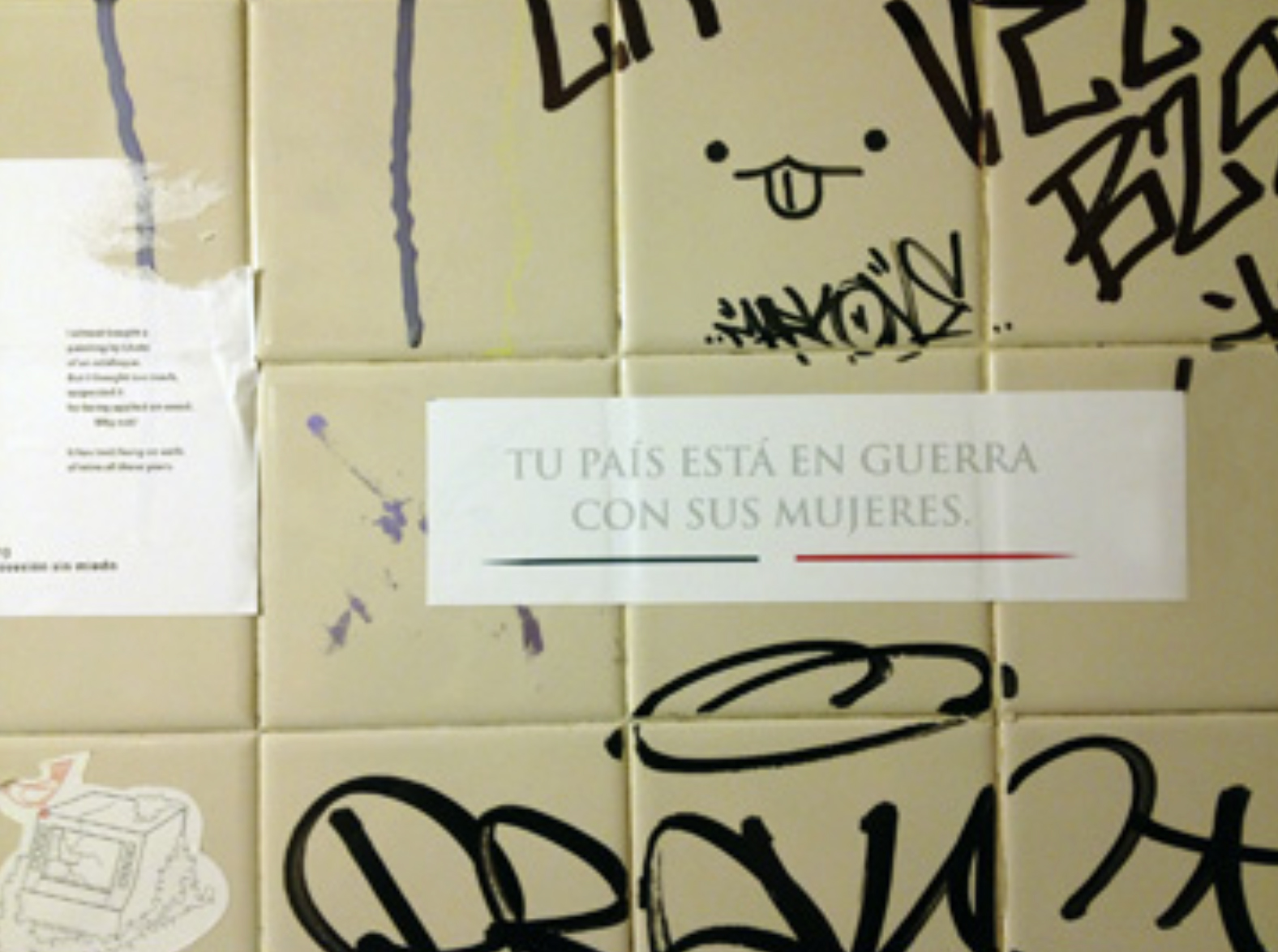 Detail of wall tiled with off-white square tiles, which has been covered with black graffiti, stickers, and a message reading "Tu País Esta en Guerra / Con Sus Mujeres"