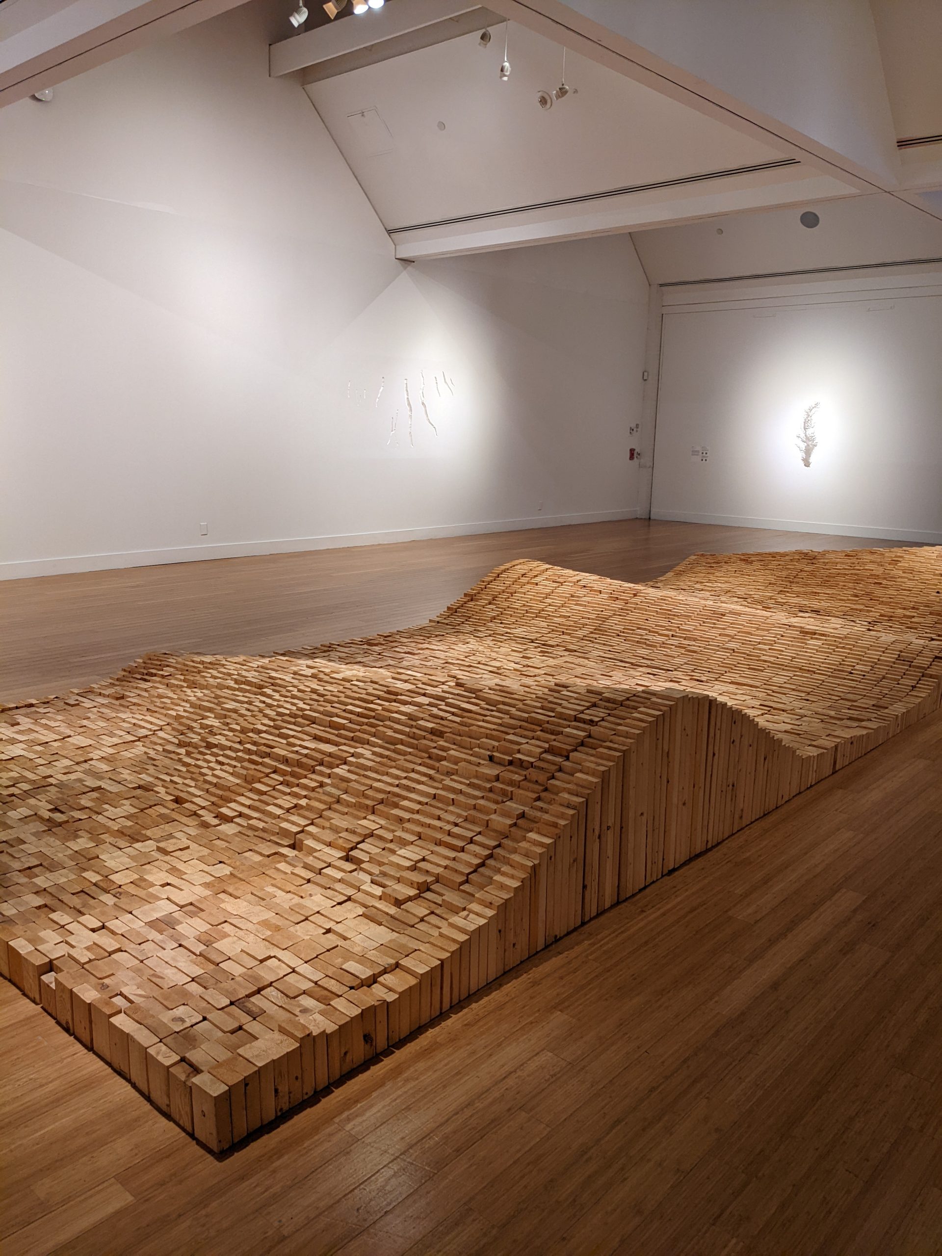View of a gallery interior with a sculpture made of sections of unfinished 2 x 4 wood timbers cut at different heights to create a wave-length effect. The sculpture is set directly on the hardwood floor.