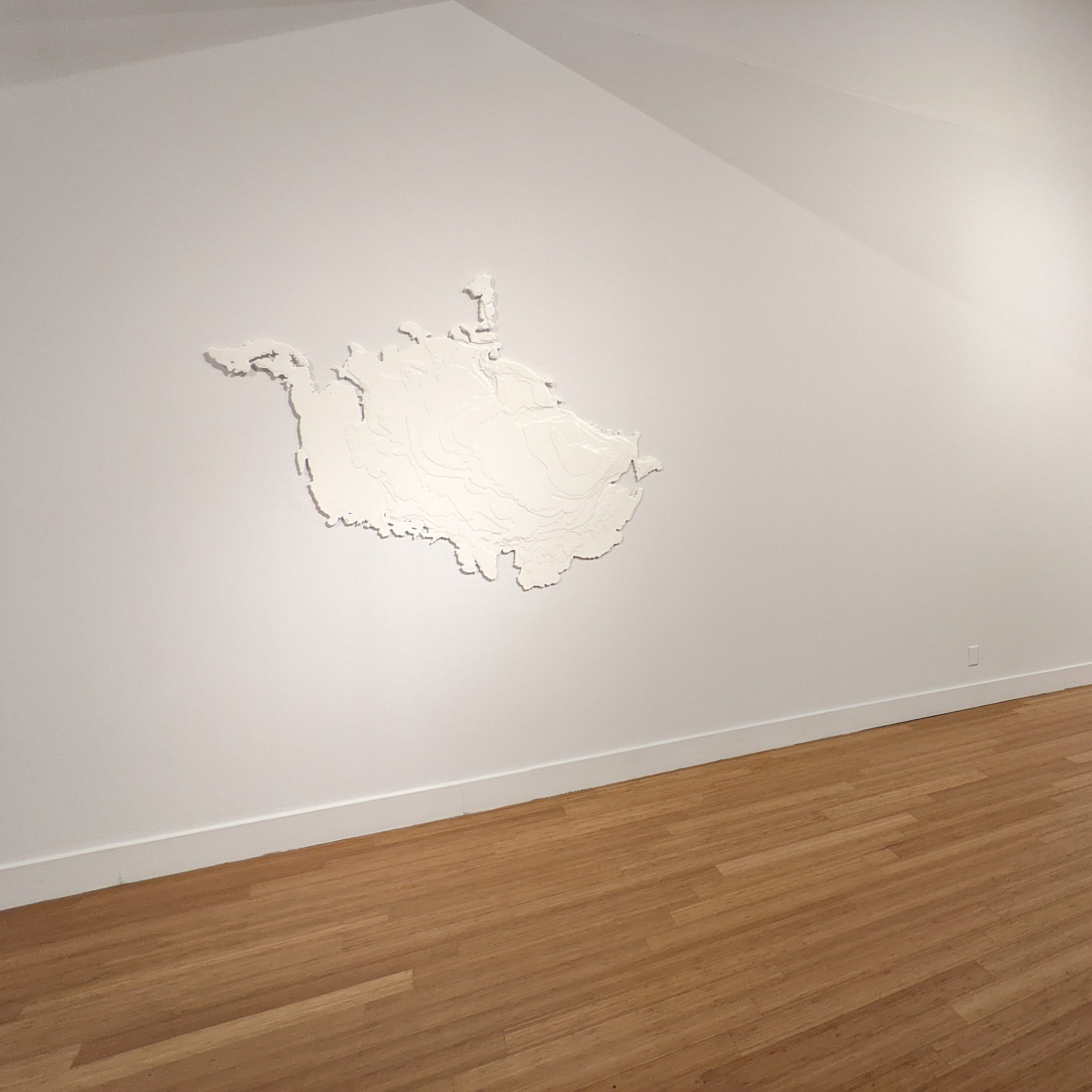 View of museum gallery interior with a white map-like form hanging on a white wall, above a hardwood floor