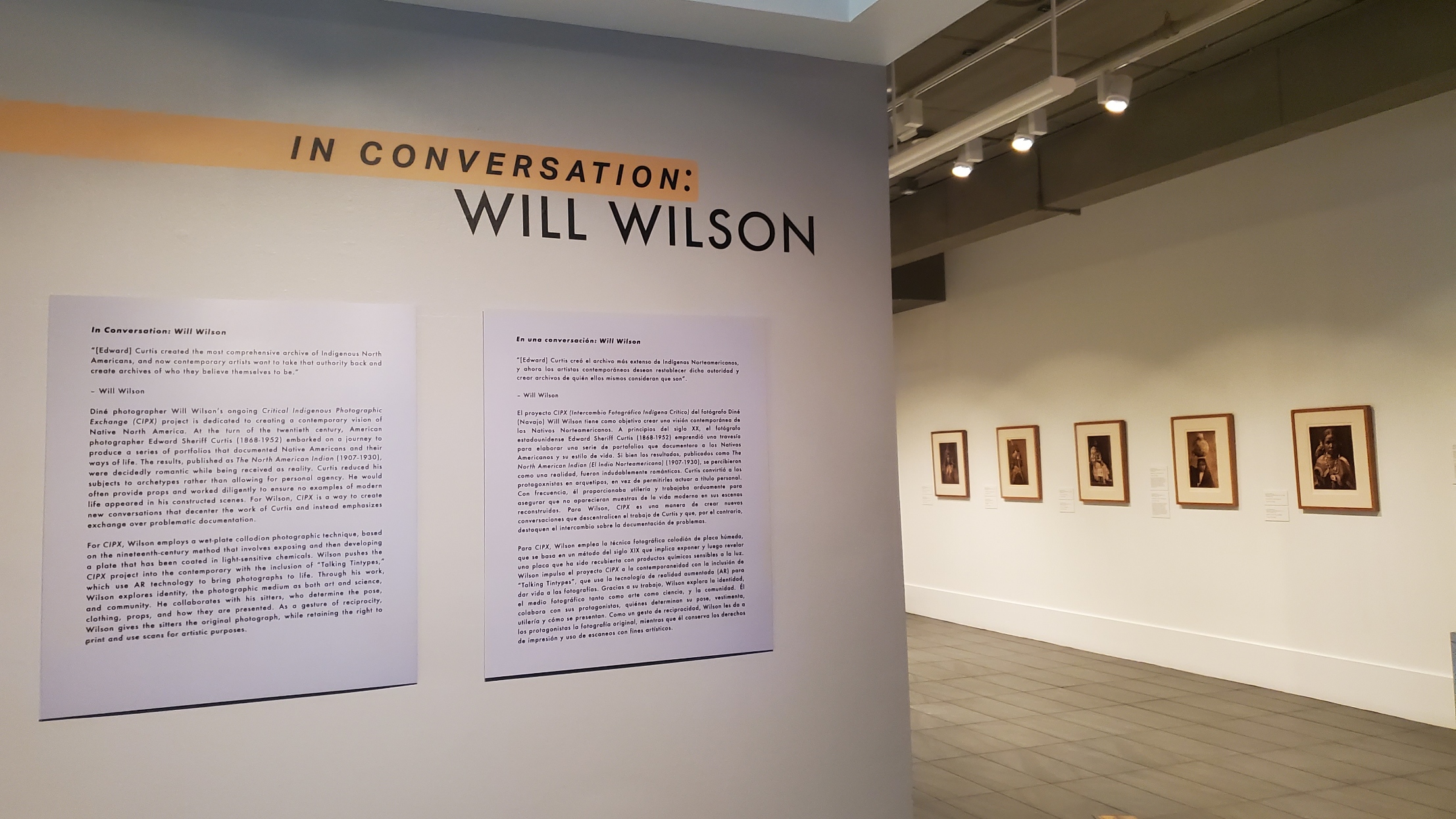 View of a museum interior showing the opening wall for the exhibition "In Conversation: Will Wilson," with two text panels side by side. Just beyond, five sepia-toned photographs are visible on an adjacent wall.