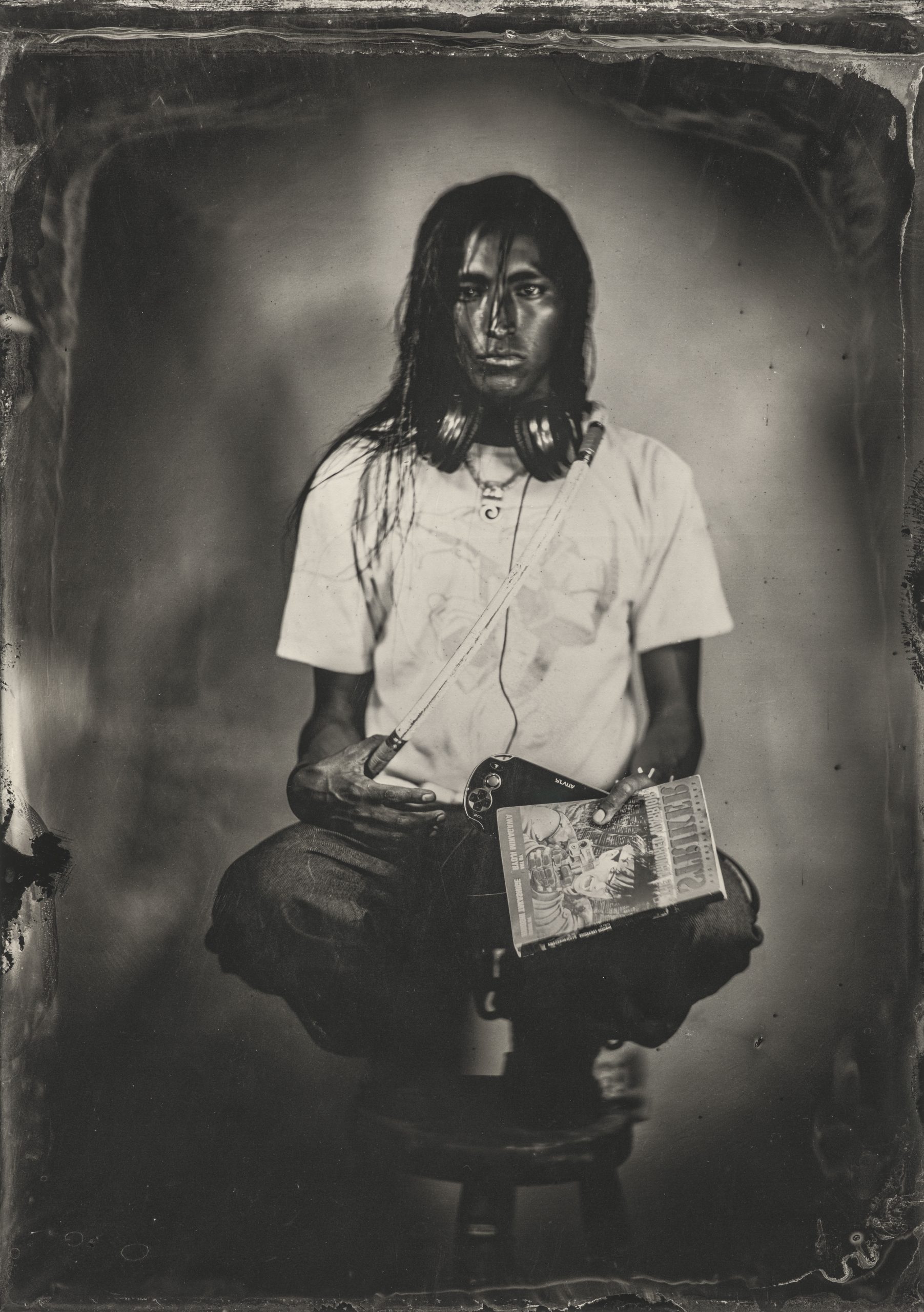 Black-and=white photograph of an Indigenous American person crouched on a stook, wearing a short-sleeved white shirt and holding a magazine in one hand.