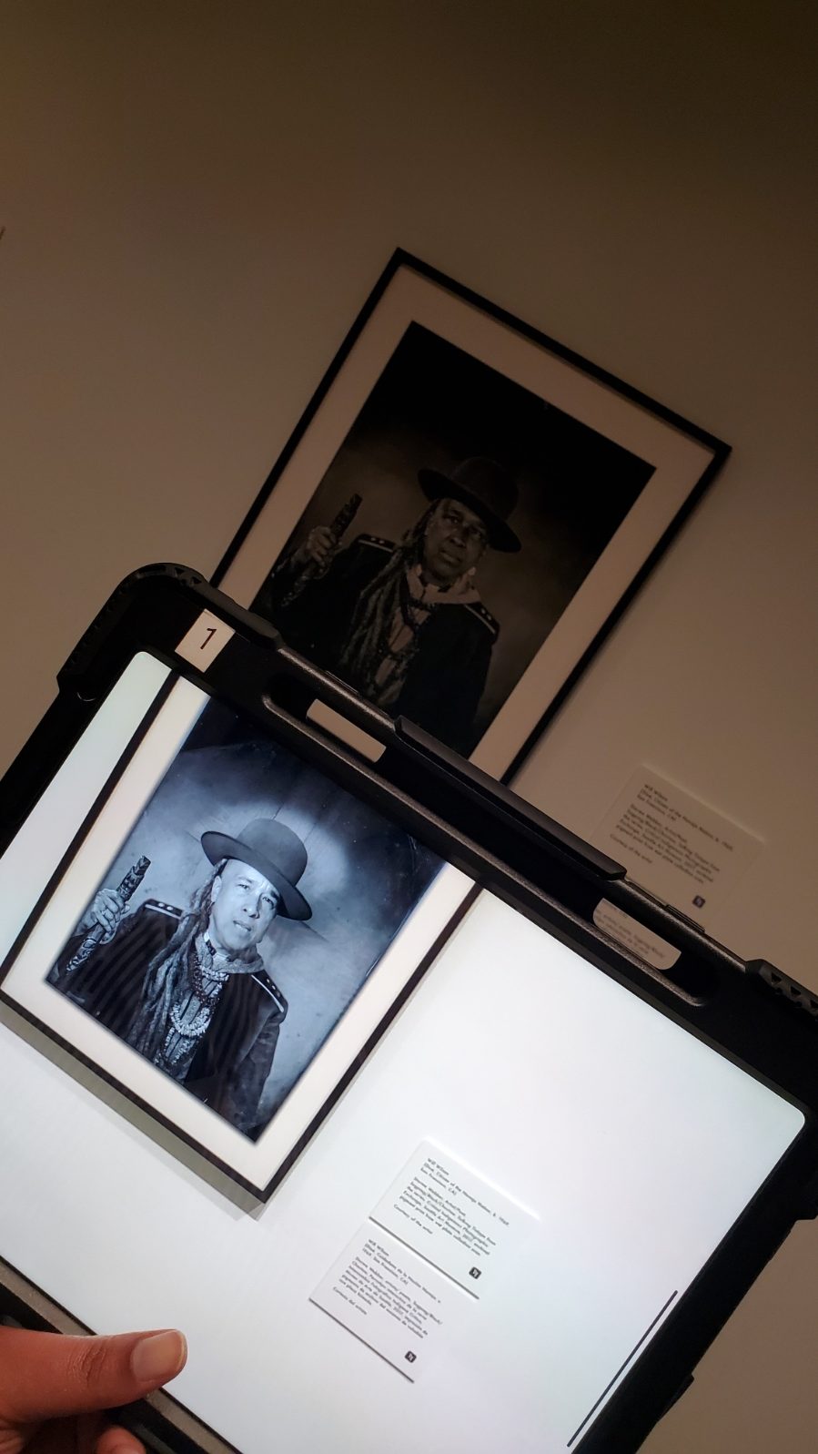View of a hand holding a computer tablet showing a black-and-white photograph of an Indigenous American wearing a broad-brimmed hat; just beyond, a similar photographed, framed and hung on a gallery wall, is visible.