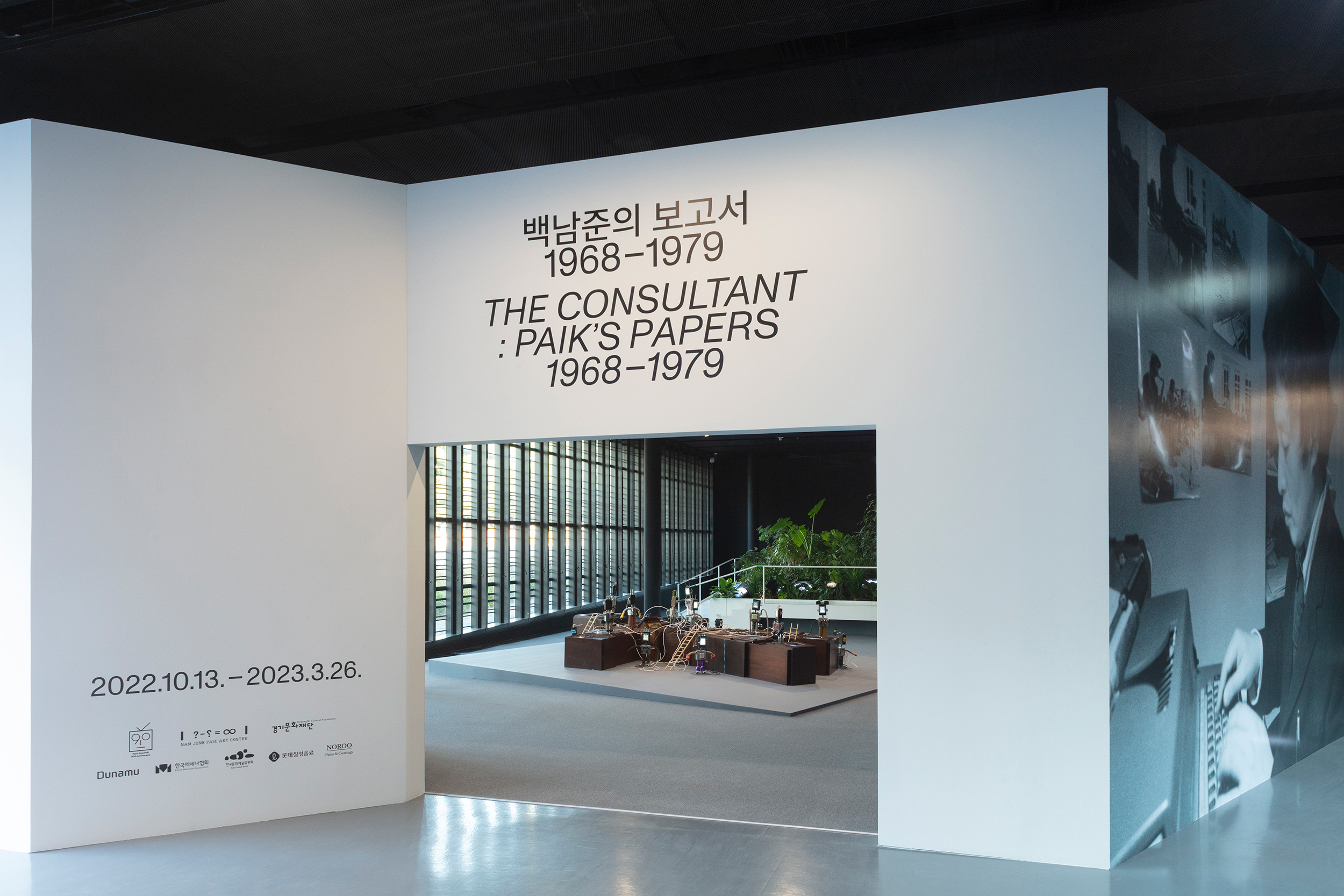 View of museum gallery with opening wall for the exhibition "The Consultant: Paik's Papers 1968–1979," with title text in both English and Korean. On an adjacent wall is a large black-and-white photograph of the artist at work; through an opening in the title wall an art installation and some greenery are visible.