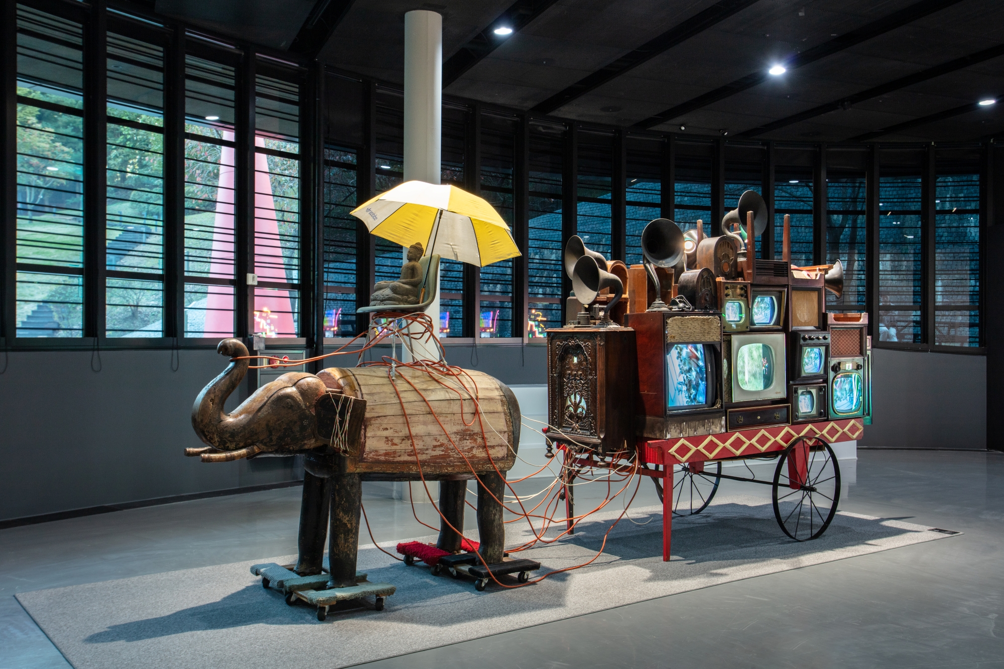 Installation artwork in the form of a cart drawn by an elephant, whose four legs rest on dollies with casters. A human figure seated in a chair and protected by an umbrella is on top of the elephant. The car the elephant draws carries multiple television monitors and old-fashioned gramophones. 