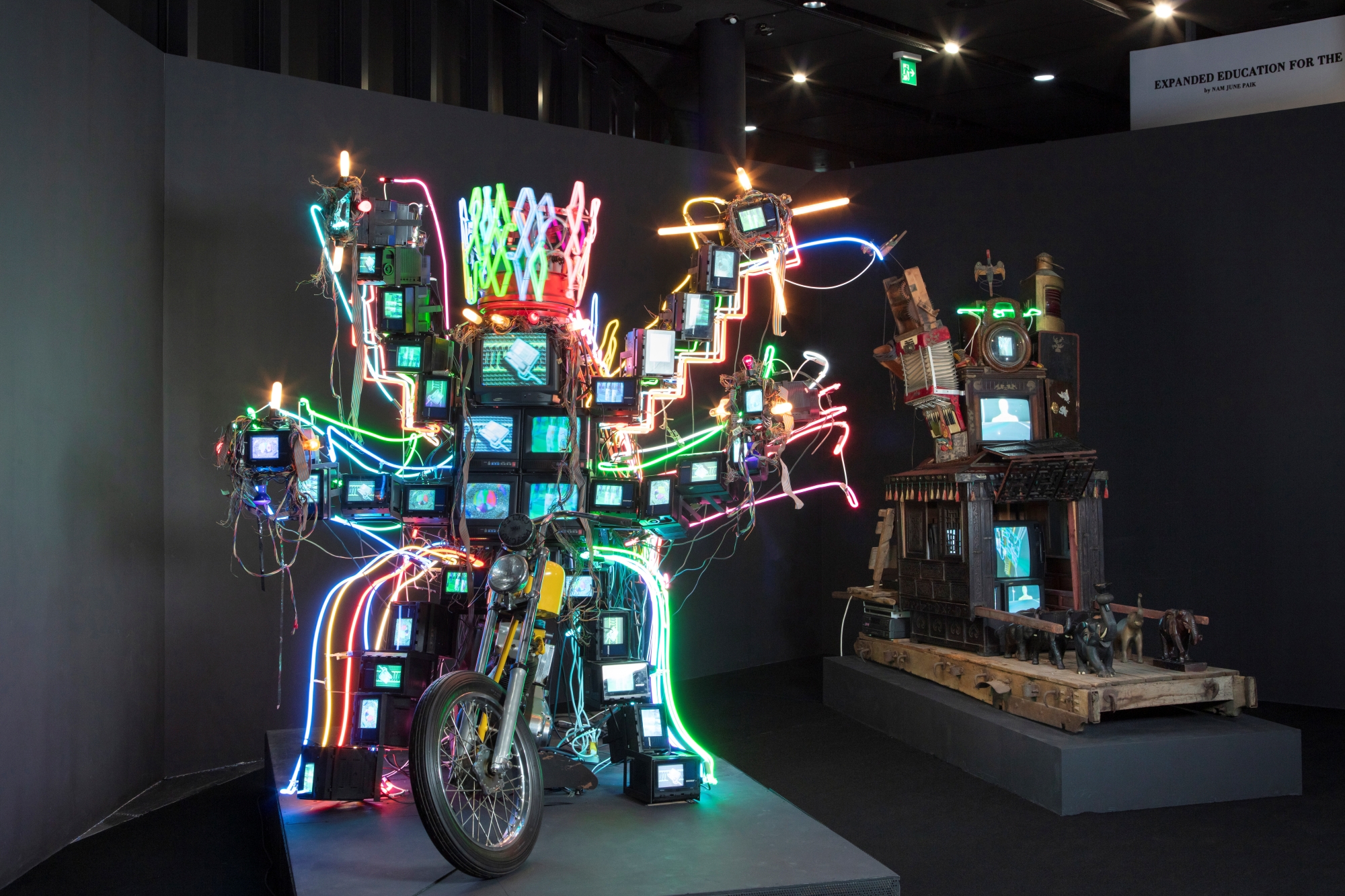 Two sculptural installation artworks side by side, both of which use multiple television monitors. The work on the right also uses colored neon and the basic form of a motorcycle.