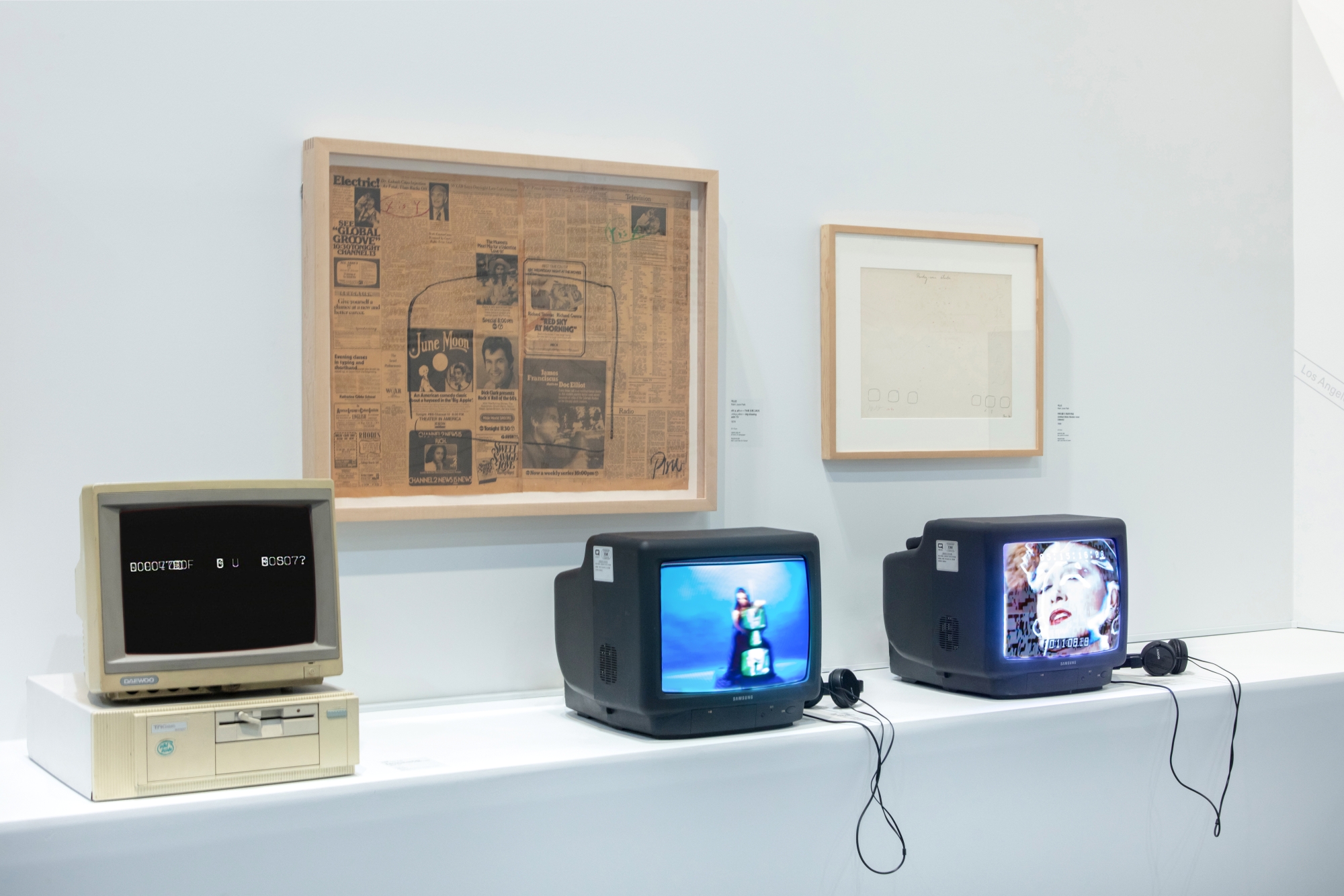 White shelf in a museum gallery holding a 1980s-vintage computer and two tube TV monitors with headphones attached. Two framed artworks are on the wall behind them.