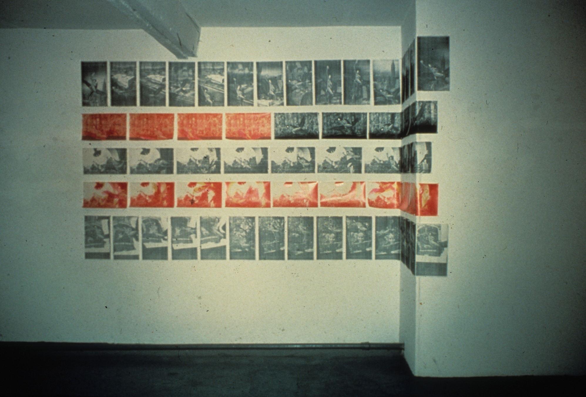 Image of the interior of a museum or gallery with a series of grayscale and orange-red compositions arranged in a grid and extending over a dogleg corner of the wall.