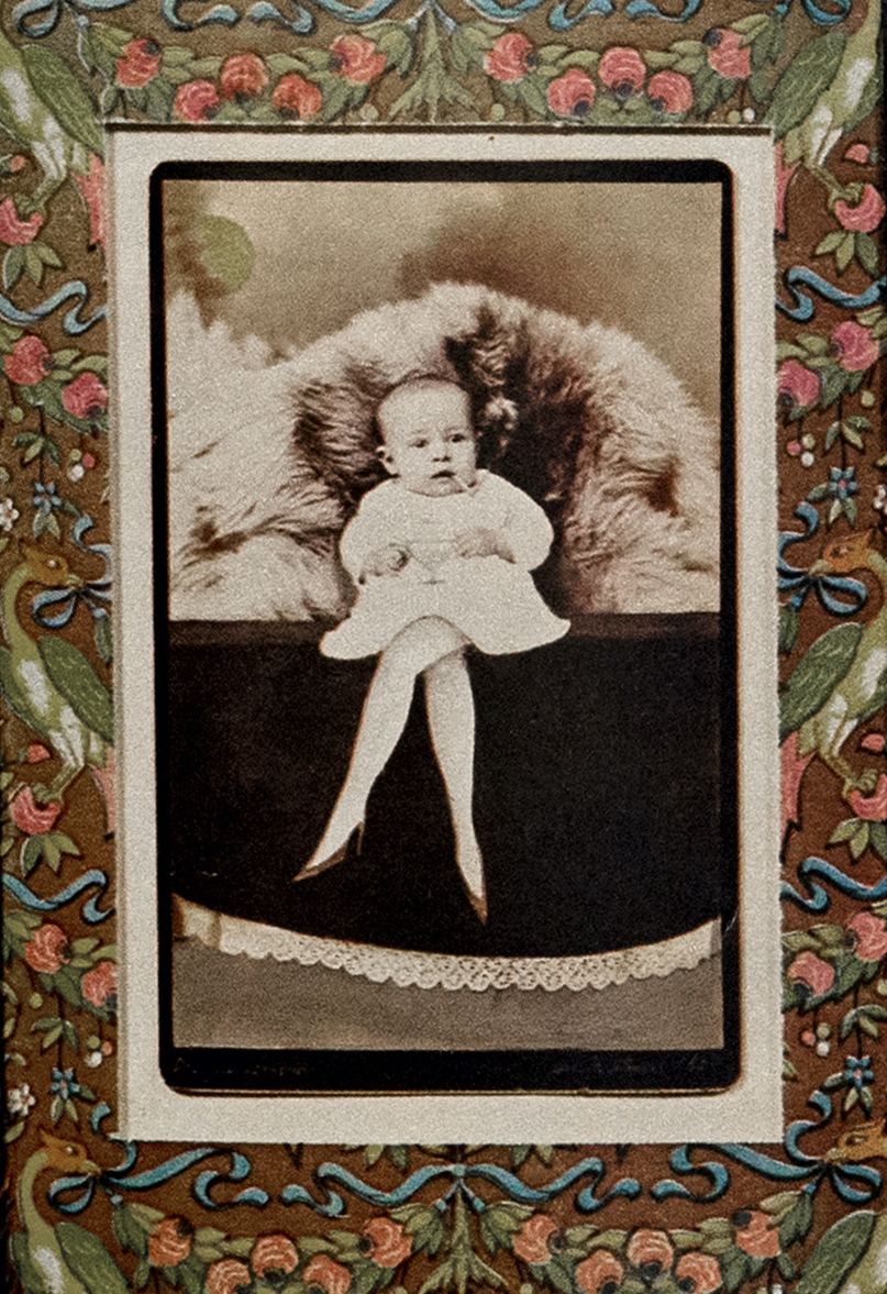 Black-and-white photograph of a baby in a white dress, altered to have unnaturally pointy feet in high heels. The baby sits on what looks like a ledge, holding a wine glass, in front of a pile of fur. The photograph has a decorative floral border.