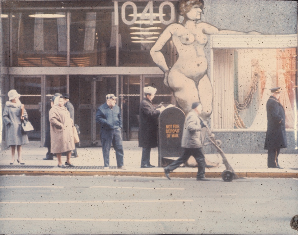 Faded color photo of a streetscape with several men and women standing on a sidewalk near a metal container reading "Not for deposit of mail." A larger-than-life cutout or painting of a nude woman rises behind them.