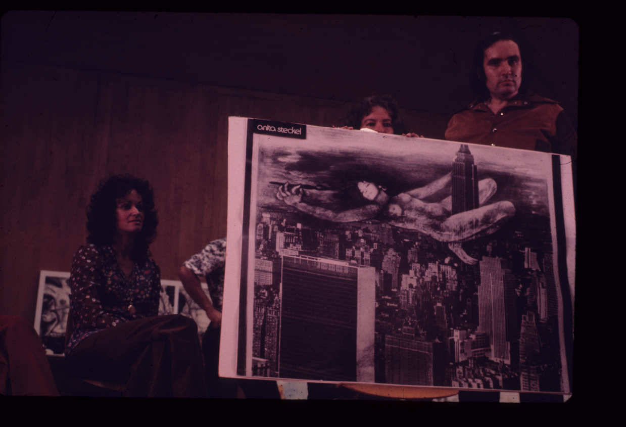 Vintage color photograph of a man and a woman holding up a black-and-white composition in which a larger-than-life-size nude woman straddles the Empire State Building like King Kong; in the upper right of the composition appear the words "anita steckel." Another woman sits to the left.