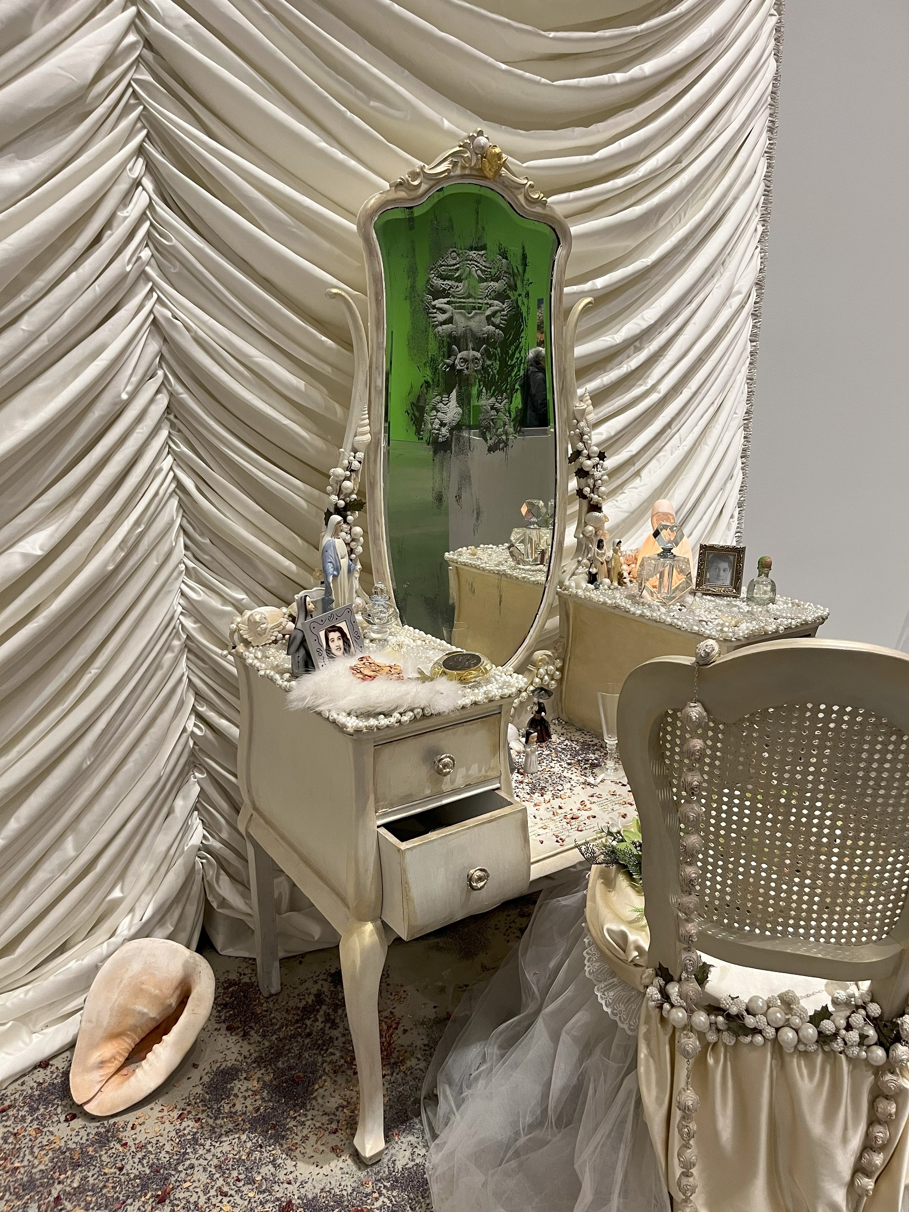 White-painted, mirror-topped vanity covered with decorative objects, in front of a ruched white curtain