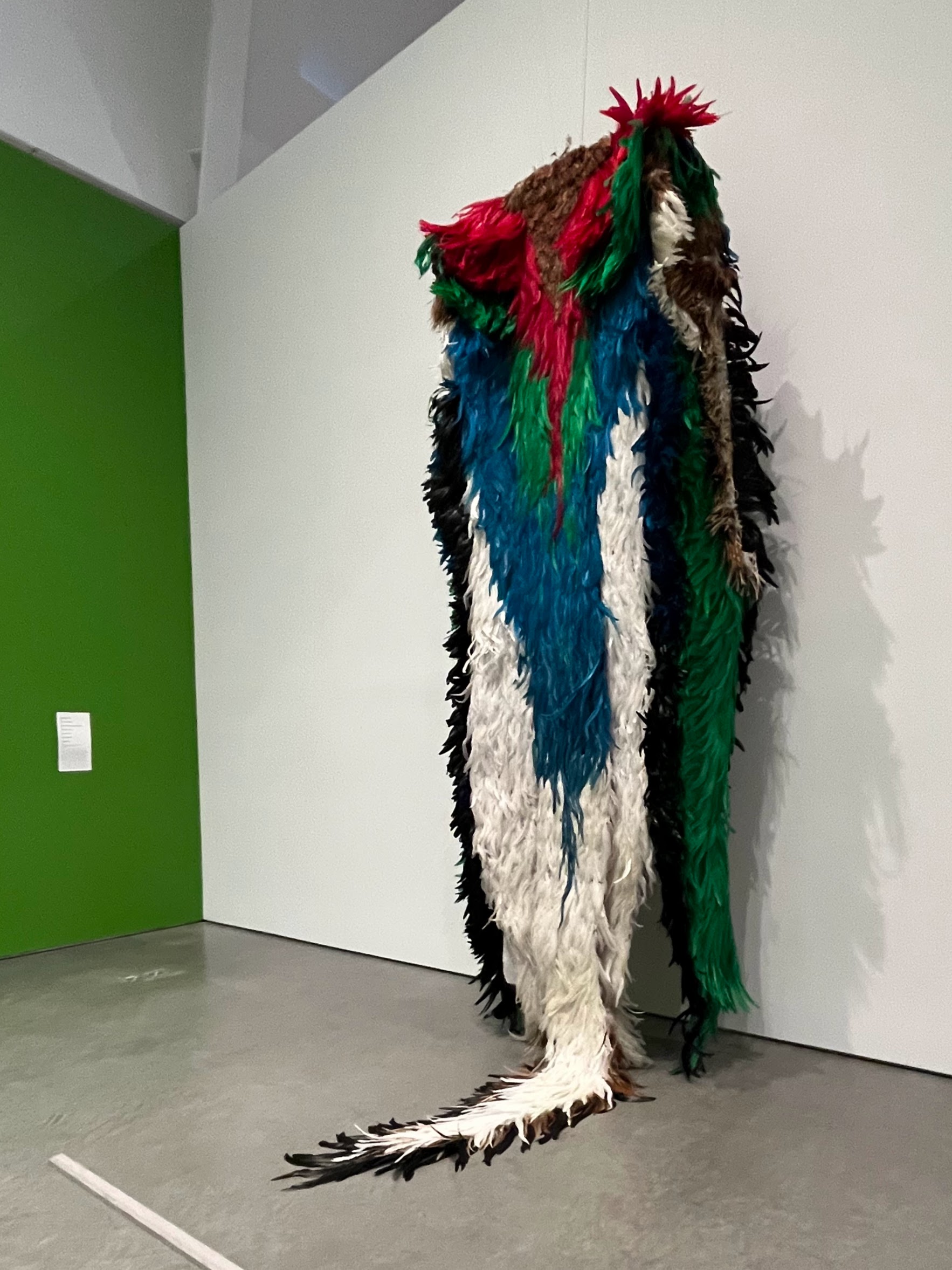 Larger-than-life-size cloak made of white, black, blue, green, and red feathers, installed in the corner of a museum gallery