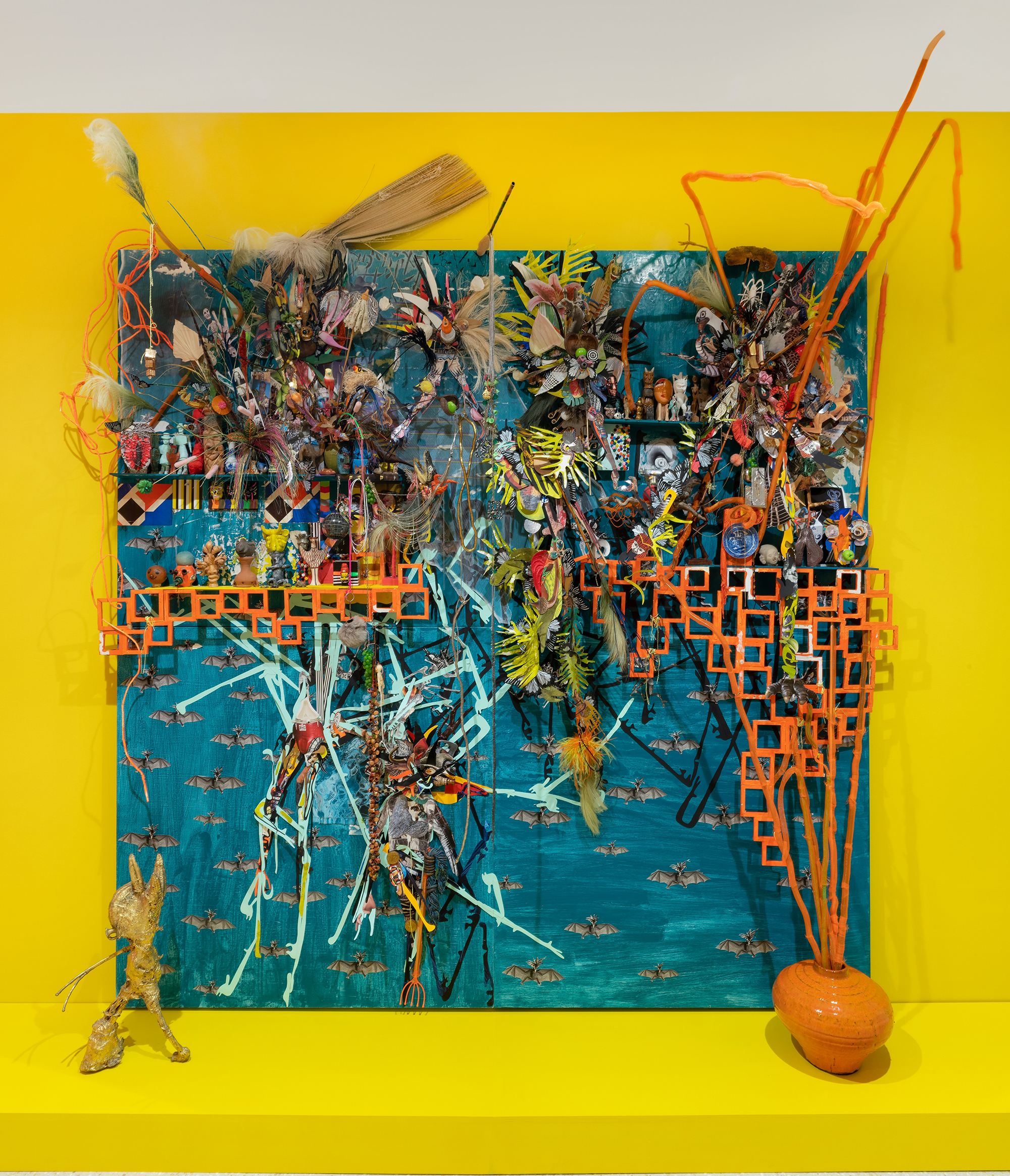 An abstract, collaged work resting on a yellow-painted plinth against a wall of the same color. The work has a blue background covered with flying bats, with collaged elements on top. A golden semifigural sculpture and an orange vase are on the plinth.