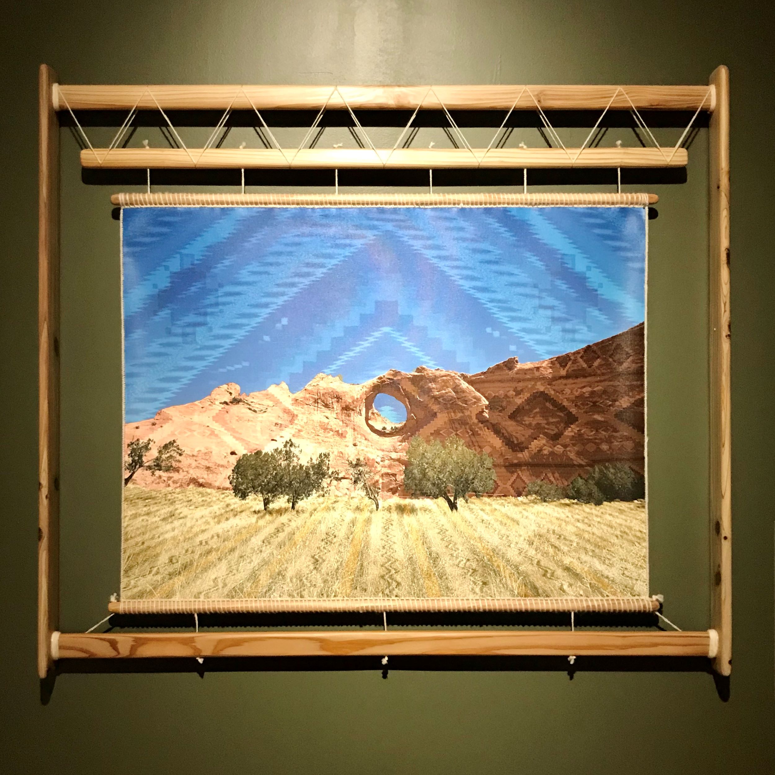 Weaving depicting a desert landscape with geometric patterns in the blue sky, framed in a loom