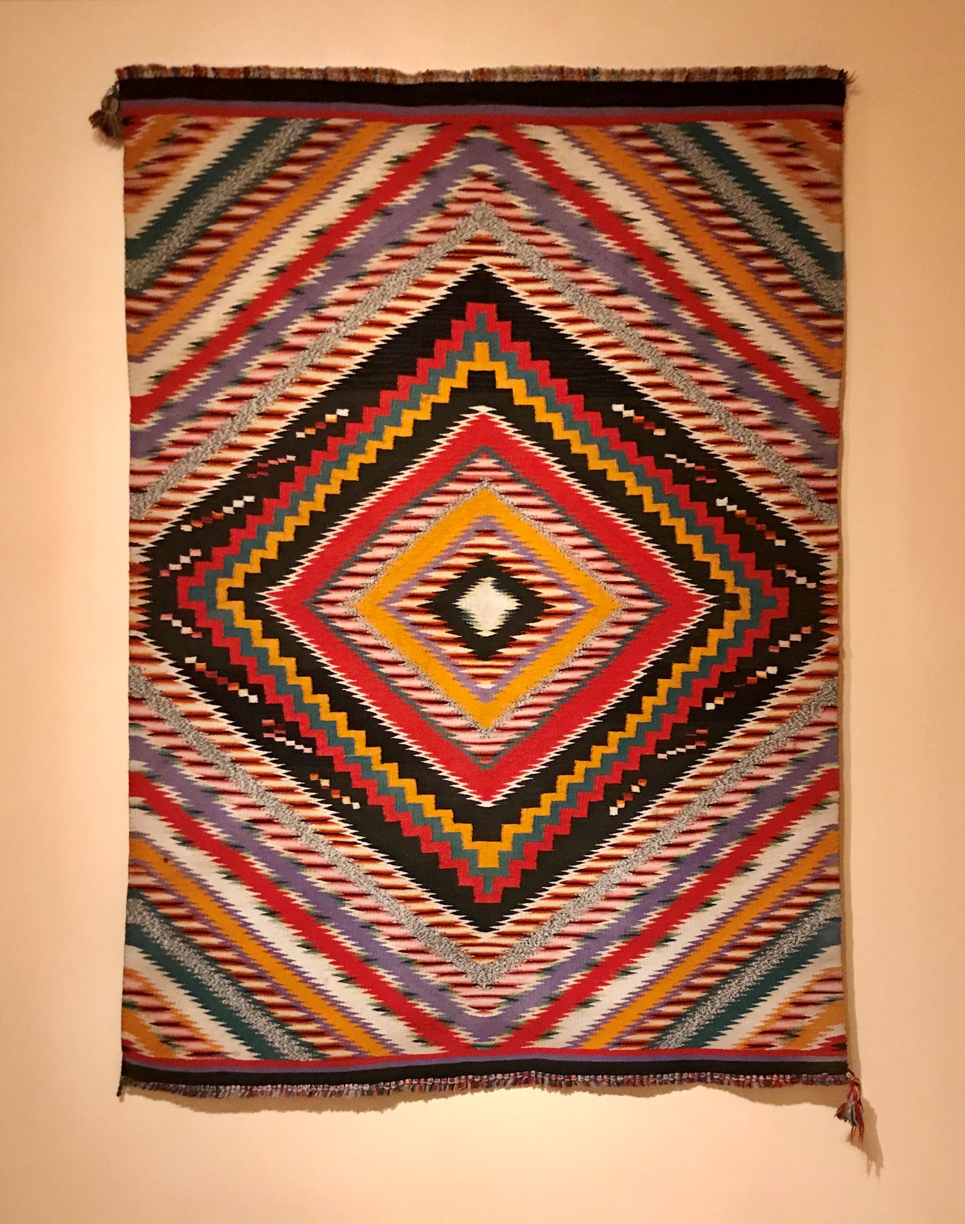 Colorful weaving hanging on a wall. The pattern is diamond-shaped and rendered primarily in black, yellow, and red.