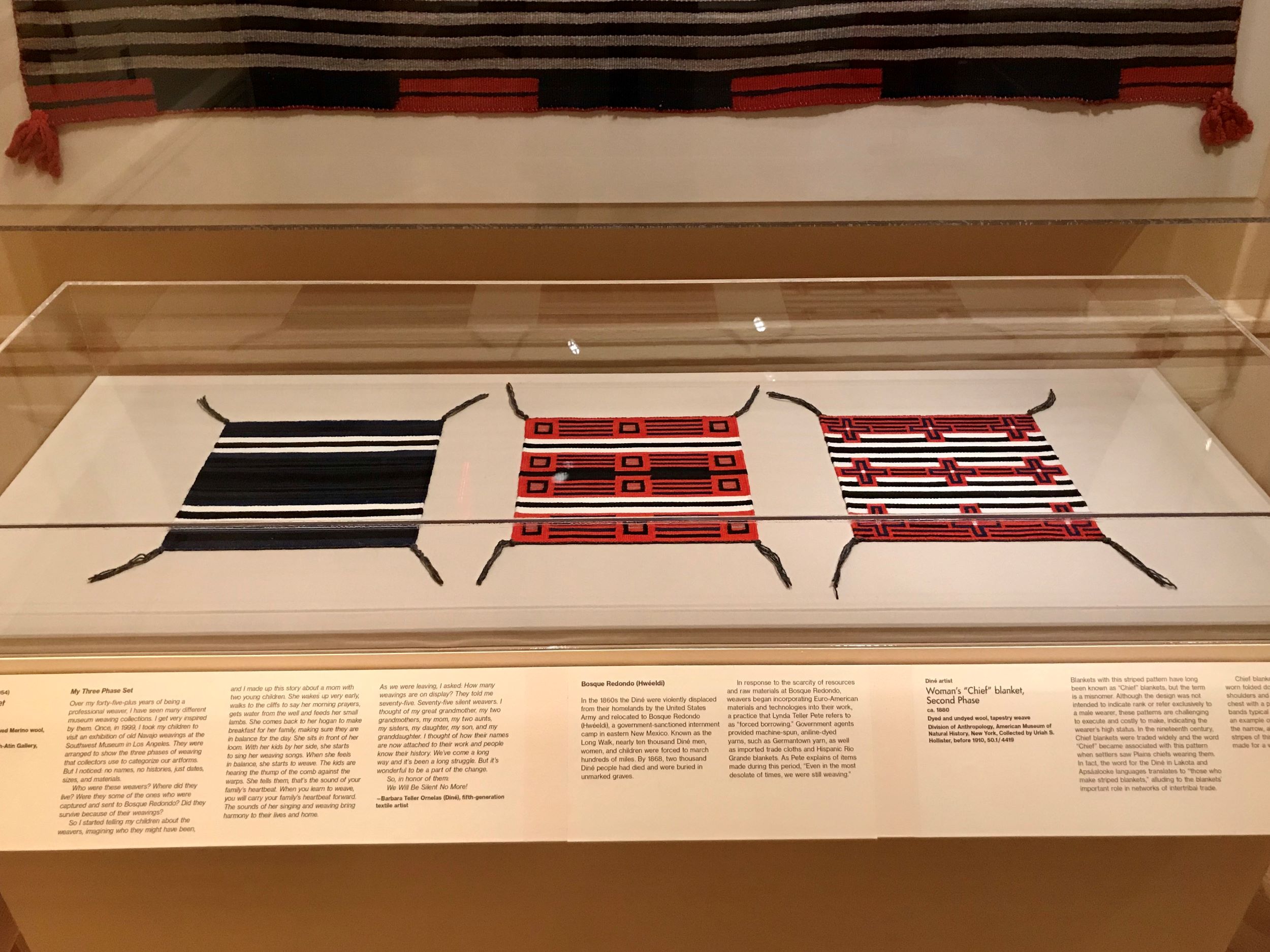 View of a glass case in a museum exhibition, with three small black, white, and red weavings inside