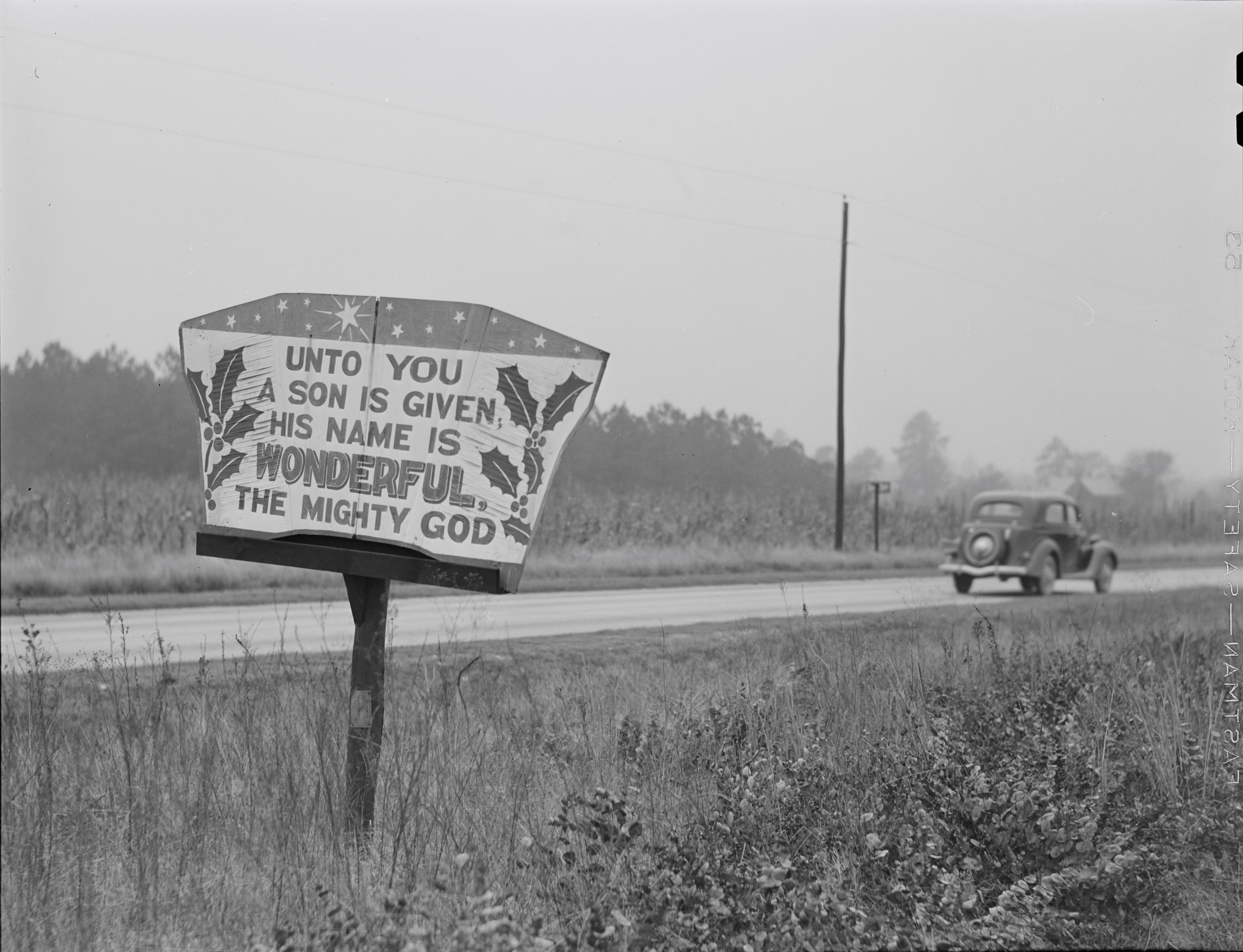 Black-and-white photograph of a rural road with a 1930s-era car driving off to the right. In the left foreground is a sign, decorated with stars and holly branches, reading "Unto you / a son is given / his name is / WONDERFUL / the might God"