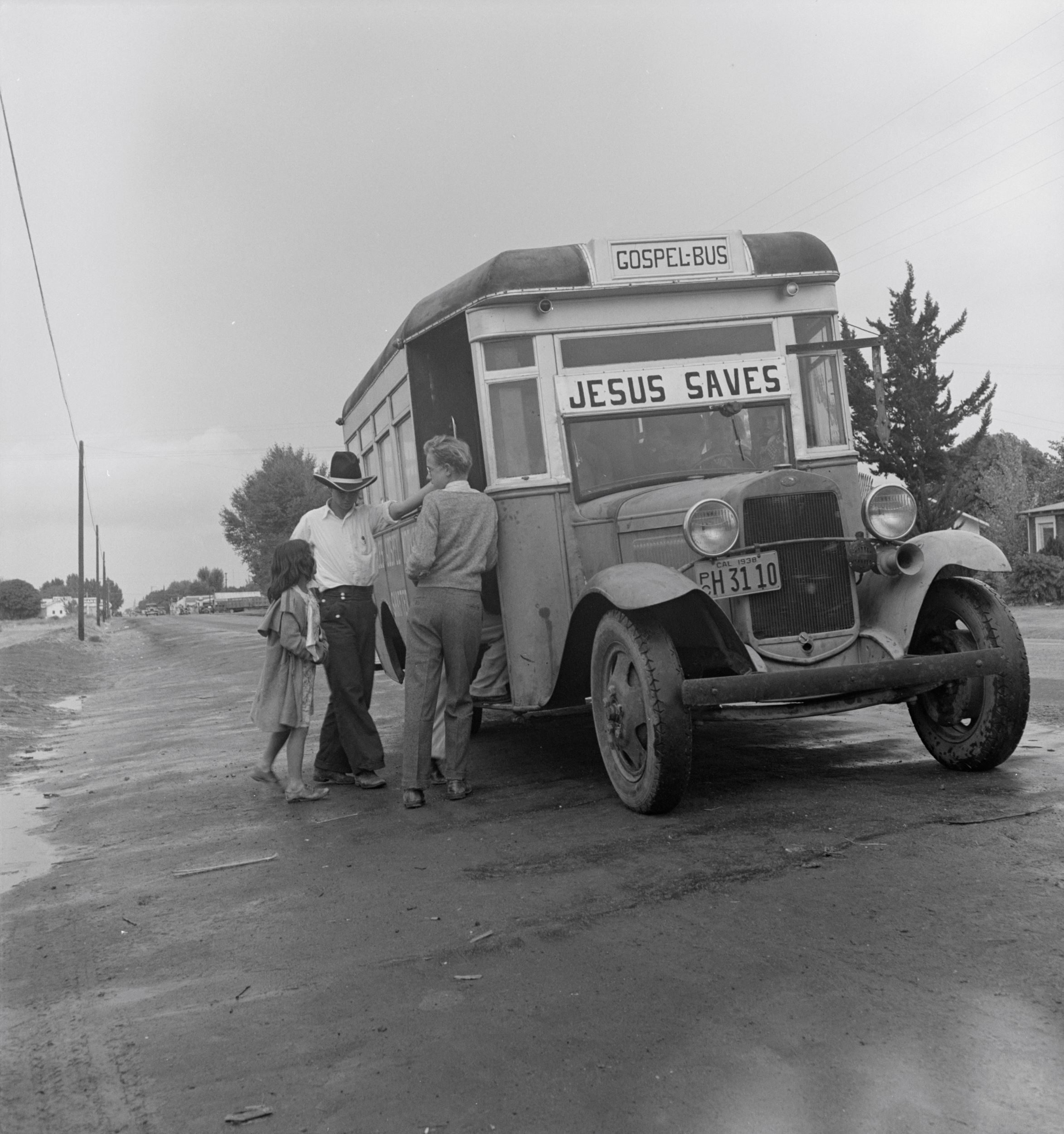 Black-and-white photograph of 1930s-era bus with a sign in the front reading "Jesus Saves." Two men and a girl linger at the door.