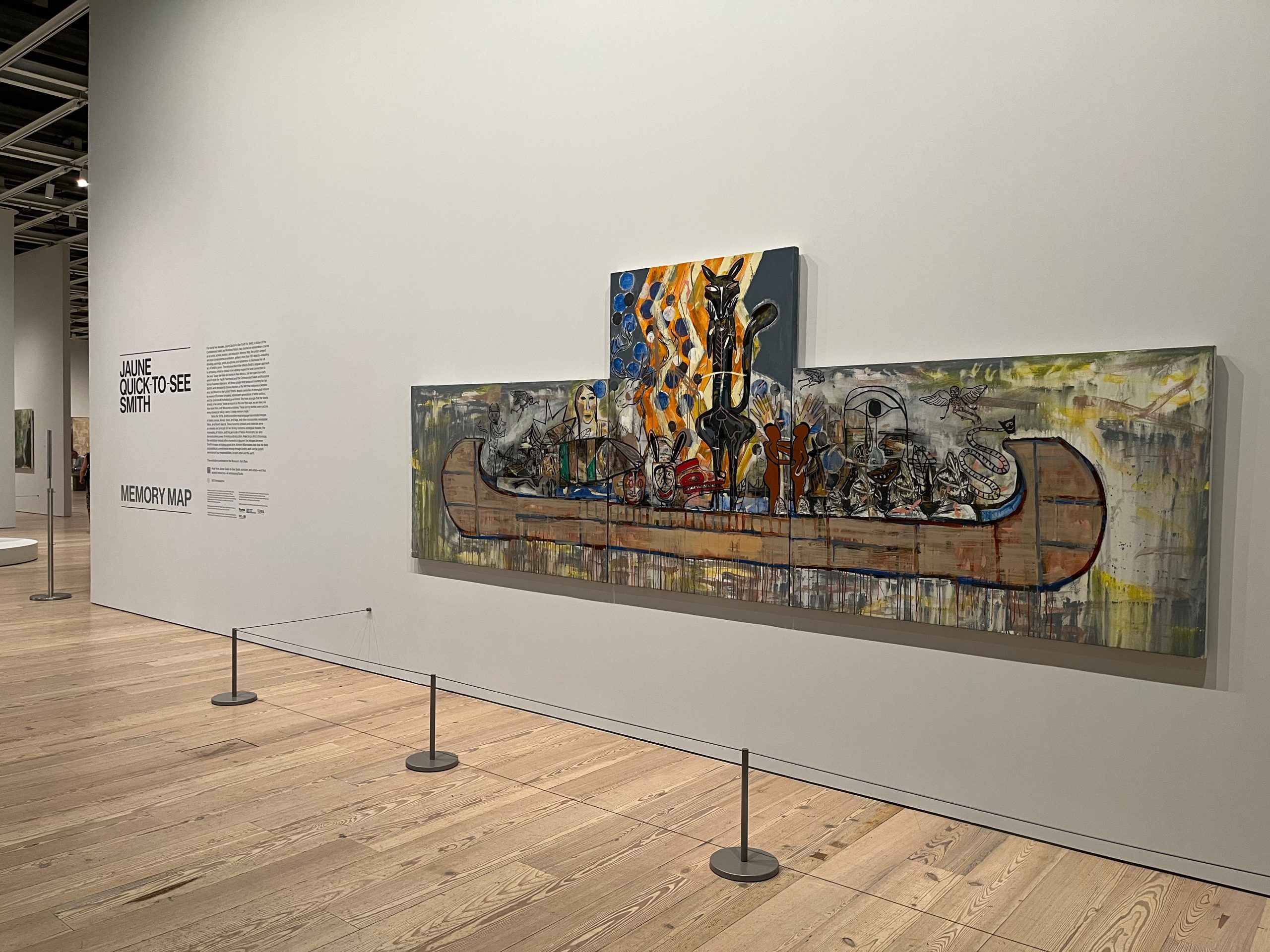 Entrance to Jaune Quick-to-See Smith exhibition, showing a diagonal gray wall in a museum interior with black text to the left and a large, colorful painting showing an abstracted canoe, piloted by a black fox in front of a bonfire.