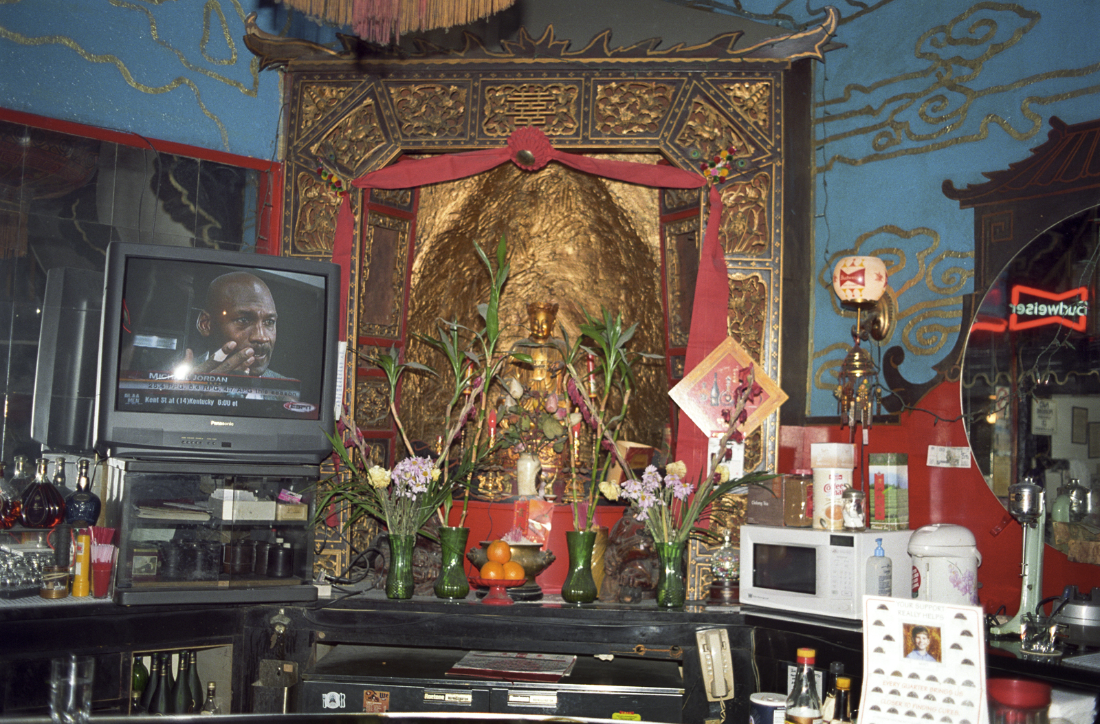 Photograph of a gold-toned Buddhist altar, draped with red, with green vases containing flowers in front. On the left is a television, showing the face of a dark-skinned man; on the right is a round mirror showing the reflection of a neon Budweiser sign.