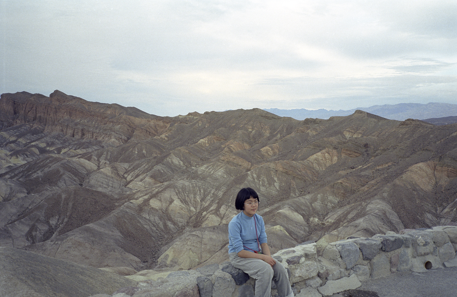 Color photograph of a young East Asian girl wearing a blue shirt, seated on a rock wall in front of a hilly desert landscape