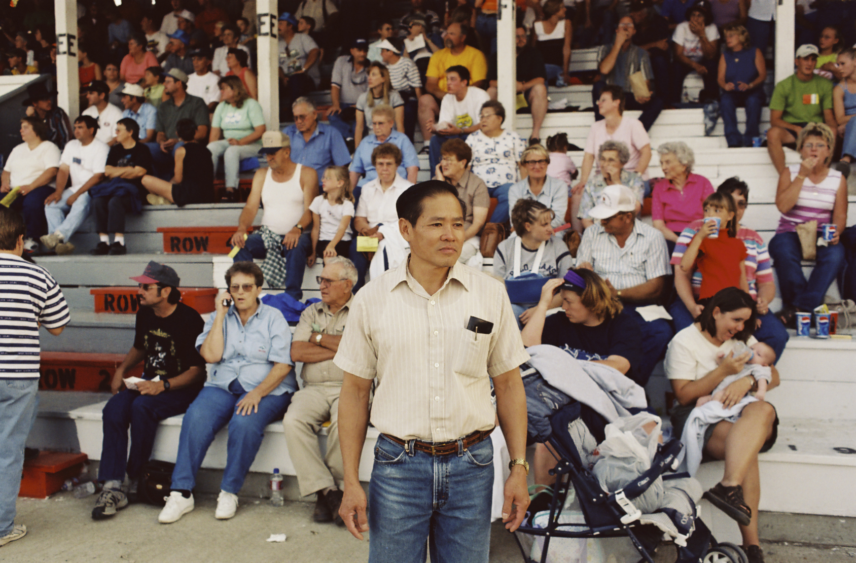 Color photograph of an East Asian man in a short-sleeved dress shirt and jeans, standing in front of a mostly seated crowd of predominantly white people.