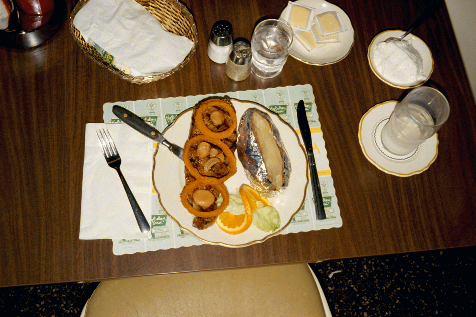 Color photograph showing a restaurant meal from above, including a paper placemat, basket of napkins, butter pats, and salt and pepper shakers