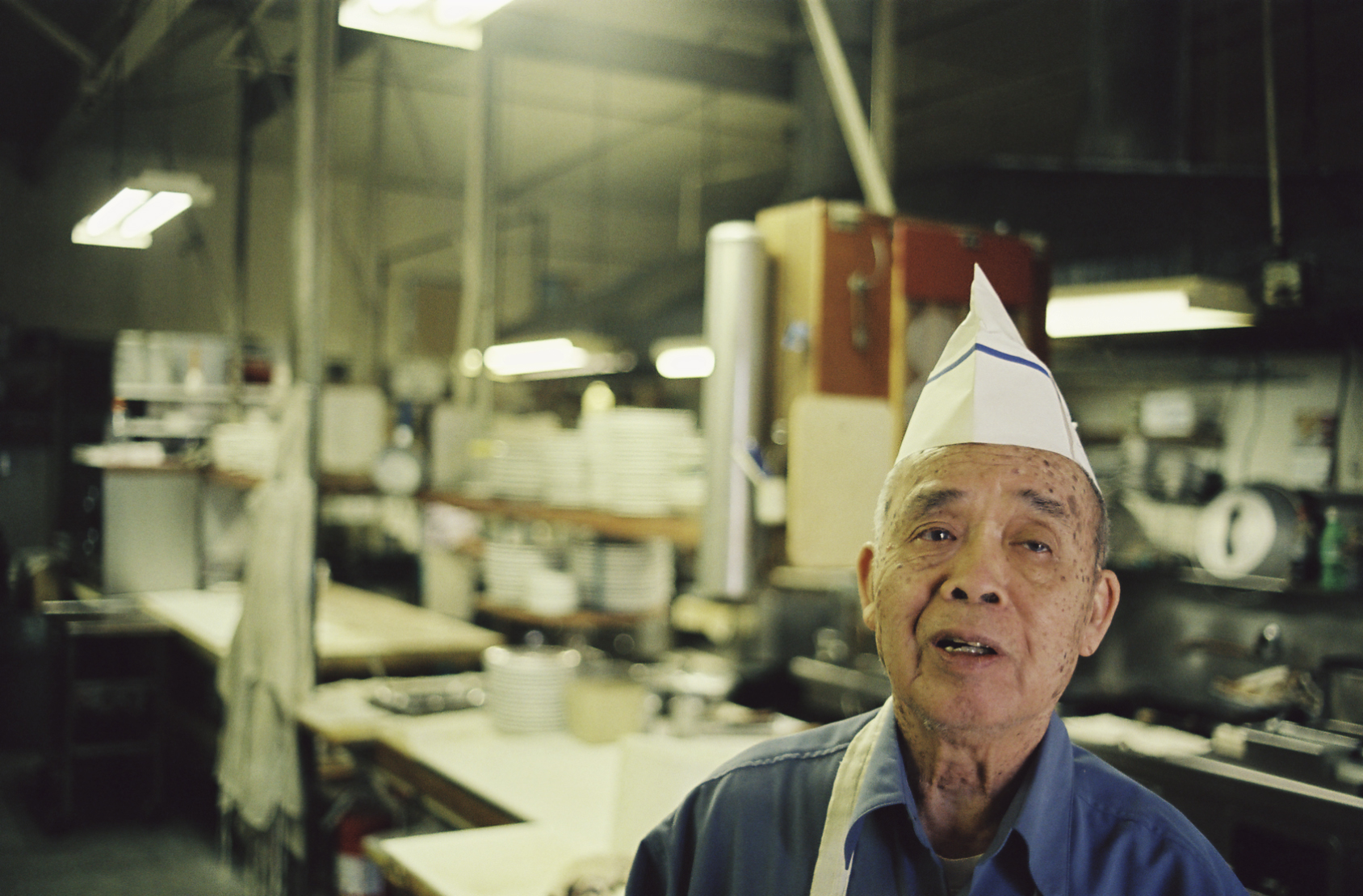 Color photograph of an East Asian man in a blue shirt, wearing a paper cap, with a soft-focus industrial kitchen behind him.