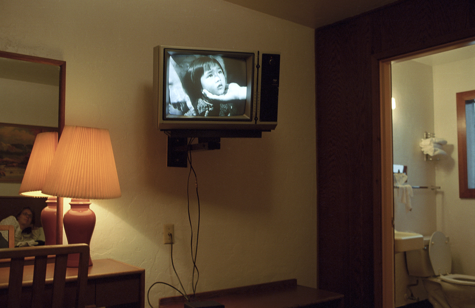 Color photograph of the interior of a room, possibly a hotel room, with a mounted black-and-white televison showing the face of a young East Asian girl. To the left is a table, chair, lamp, and mirror showing the reflection of someone lying on a bed. To the right is an open door leading to a bathroom.