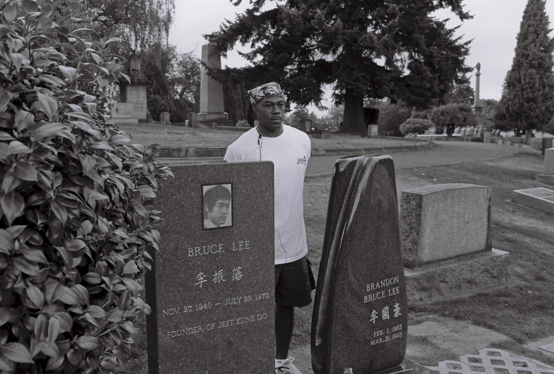 Black-and-white photograph of a dark-skinned young man wearing a white t-shirt and a bandanna, standing between two headstones that read "Bruce Lee" (on the left) and "Brandon Bruce Lee" (on the right).
