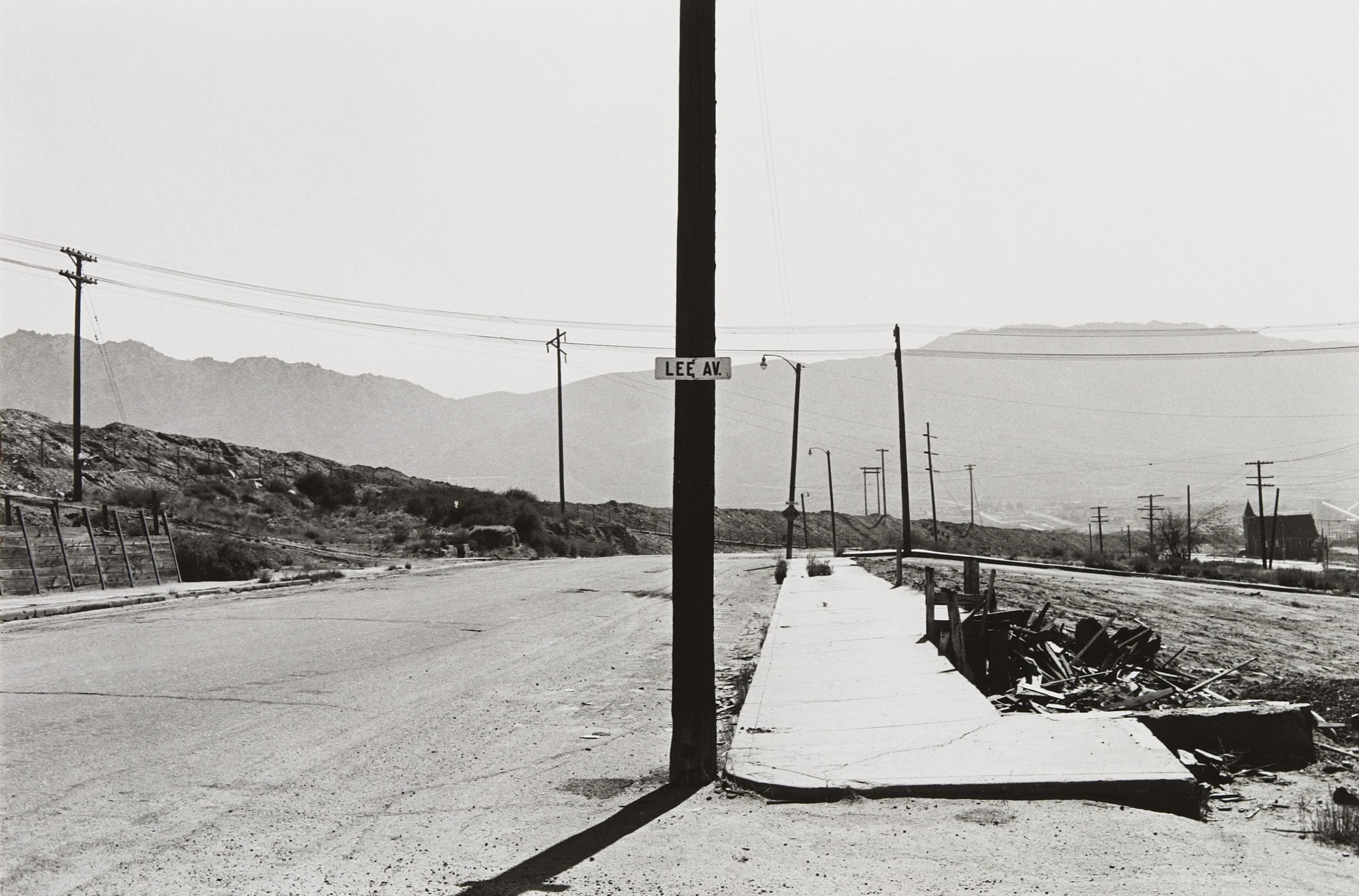 Black and white photograph of a semi-industrial rural road, bordered by telephone poles, with low hills beyond. A street sign on the centermost pole reads "Lee Av."